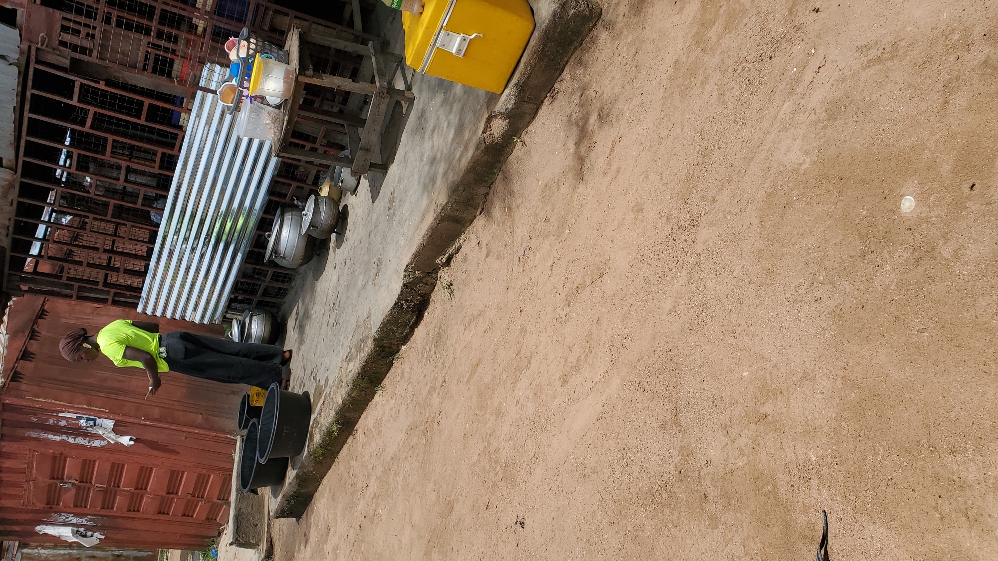 The image depicts an outdoor scene likely in Jalingo, Nigeria, with a focus on a woman standing by a collection of large black basins. The woman, wearing a bright green shirt and dark pants, is holding a mobile phone. Behind her, several metal cooking pots are visible, along with a corrugated metal sheet structure. To the right, a yellow cooler is placed on the ground. The ground itself is composed of sandy soil and a paved pathway. The overall environment suggests a commercial or informal vending area.