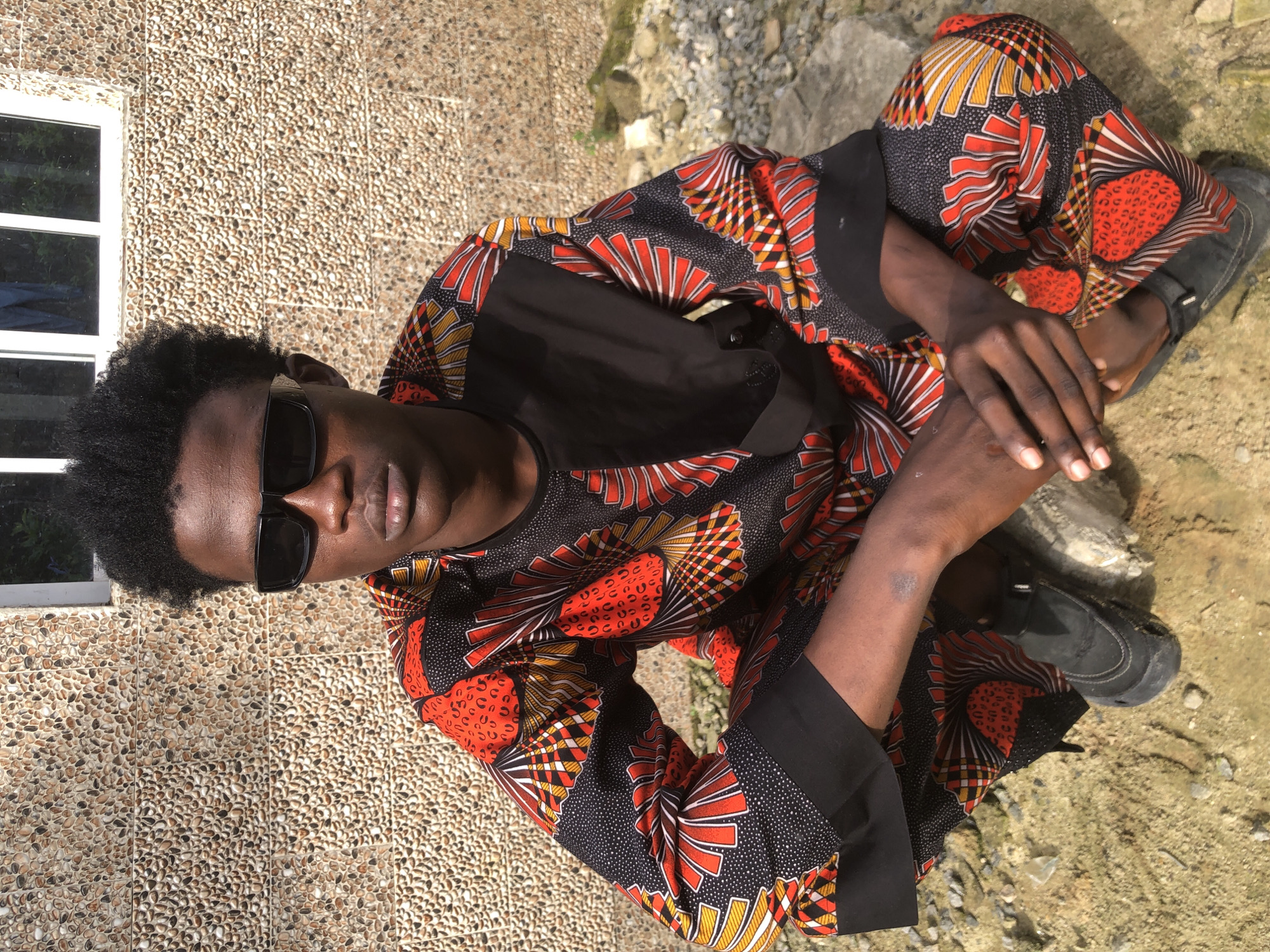 A medium shot shows a young Black male crouching outdoors in front of a textured, pebbled wall and a white-framed window. The man is wearing sunglasses, a black t-shirt, and a colorful dashiki with red, yellow, and black geometric patterns. He has dark, curly hair and his hands are clasped in front of him. He is wearing black shoes. The ground around him is sandy and rocky. The scene appears to be in Ado, Nigeria, as indicated by the presence of traditional Nigerian attire and the architectural style of the building.