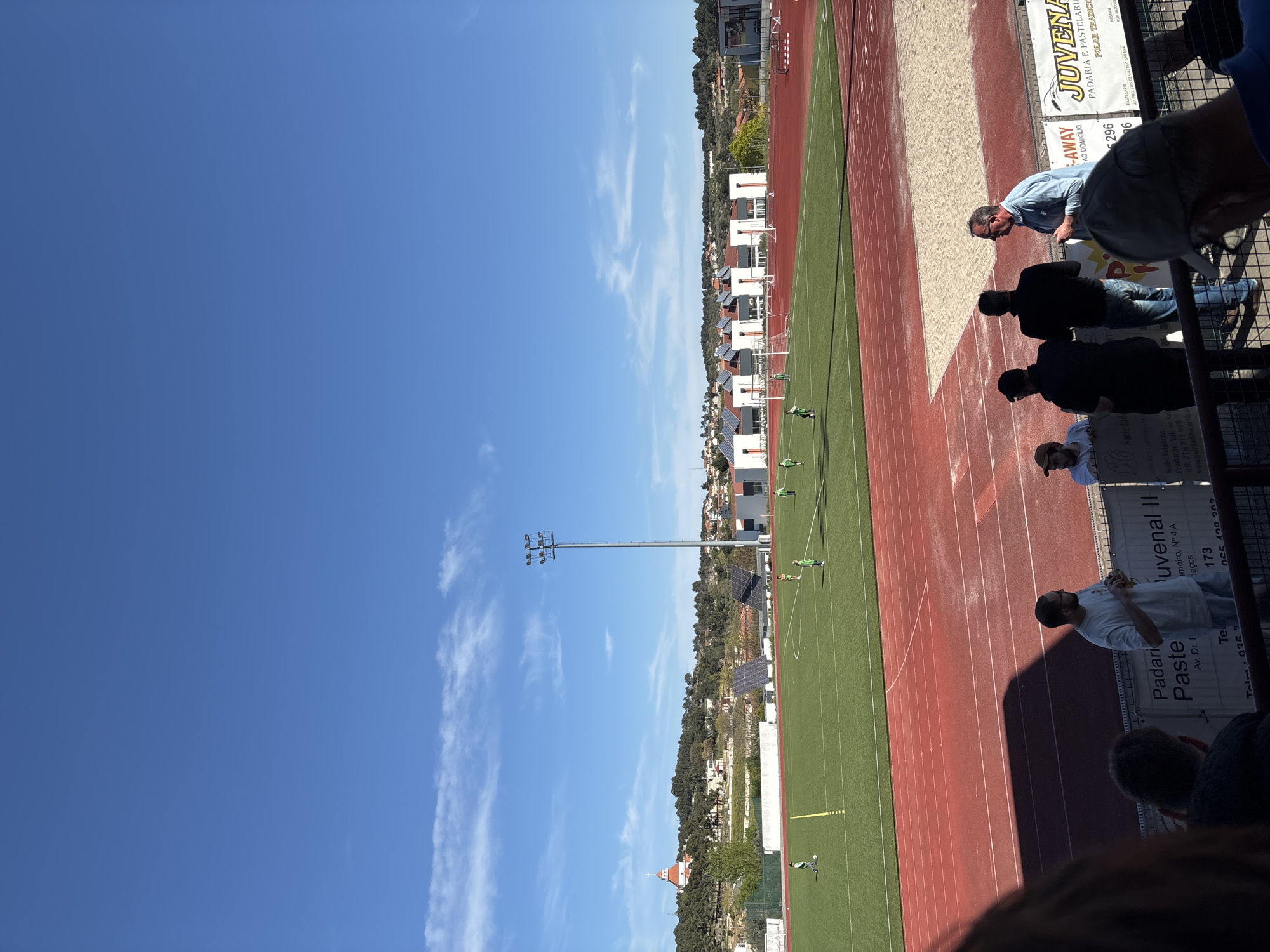 The image shows a soccer field and a track on a sunny day, viewed from the stands. A group of people watch the scene from behind a fence that is adorned with advertisement banners. The ads mention "Juvenal II," "Padari Paste," and a local "Sociedade," suggesting the field is in the municipality of Valpaços in Portugal, specifically in Valpaços, as indicated by "Sede: Valpaços" on one of the ads. Other elements in the background include houses, trees, and a lamp post, indicating an urban periphery. The sky is mostly clear with a few clouds.