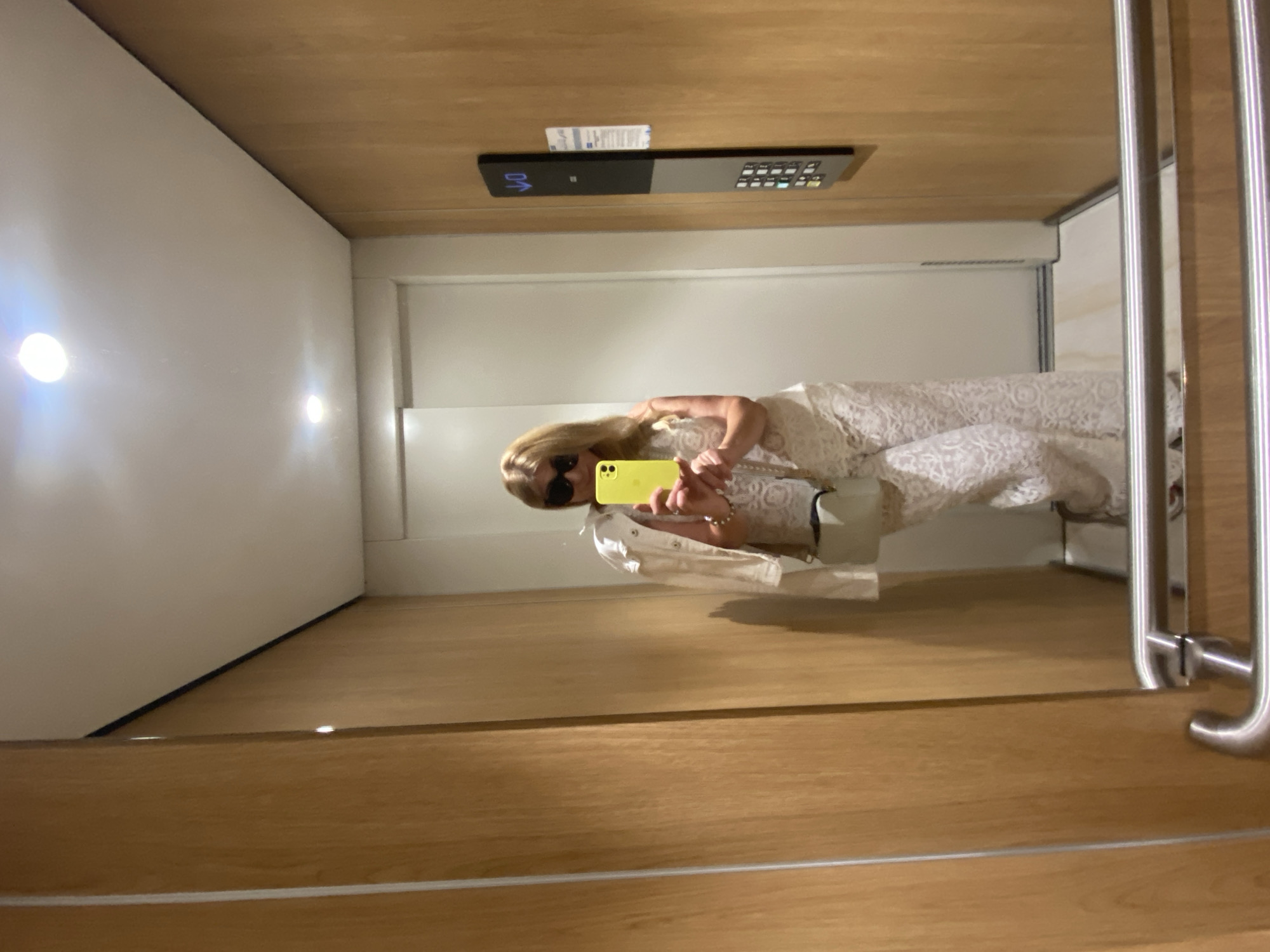 The image shows a reflection in a mirror within an elevator. The mirror reflects a person with blonde hair, wearing sunglasses, a white lace dress, and holding a yellow mobile phone. The person appears to be taking a selfie in the elevator. The elevator's interior includes white walls with two lights, a wooden ceiling with elevator controls, and metal handrails near the mirror. The location is unspecified, but based on the style and materials used, it could be in Budva, Montenegro.