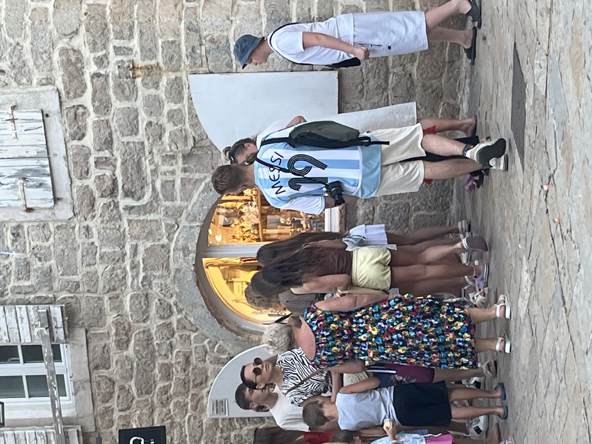 The image depicts a street scene in the old town of Budva, Montenegro, with a group of people gathered outside a shop with a stone facade. A young man in the foreground wears an Argentinian football jersey with "MESSI 19" printed on the back and is holding a camera. He is surrounded by other individuals, including women in dresses and shorts, and a young boy. The shop window displays various items, suggesting it might be a souvenir or artisan shop. The cobblestone street and weathered stone building indicate a historic urban environment.