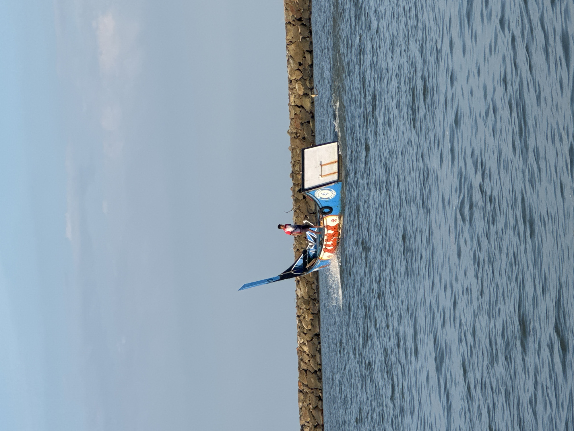 A single individual stands on a decorative, blue and orange boat as it moves through choppy, blue water. The boat has an elaborate, upturned blue stern and a white rectangular object attached to its side. The boat is creating a wake as it travels. In the background, a long, rocky breakwater stretches across the horizon. The sky is a light, hazy blue with a few wispy clouds. The scene appears to be in the coastal city of Pante Beurene, Indonesia.