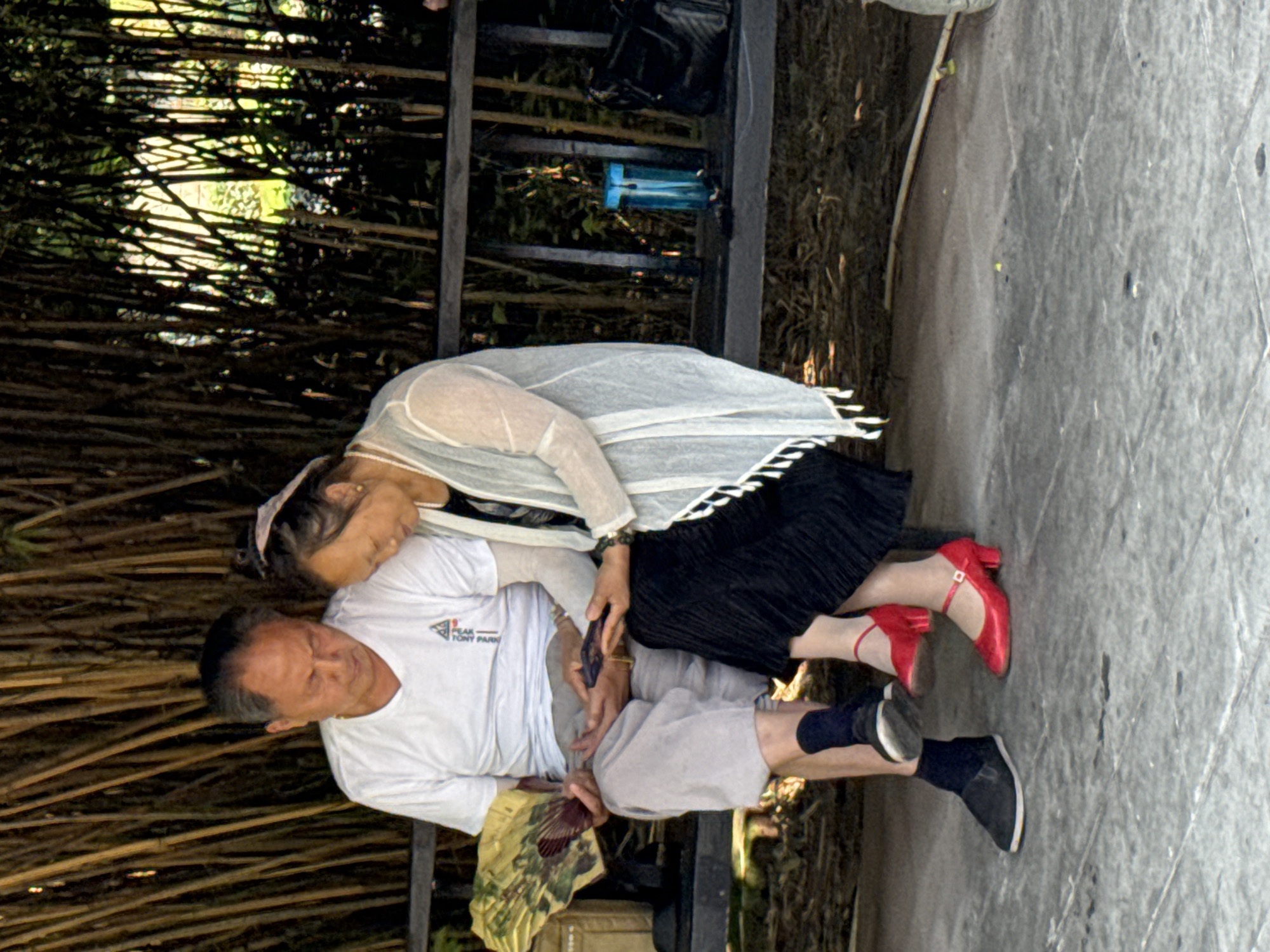 This image, rotated 90 degrees counter-clockwise, depicts an older man and woman resting together outdoors in what appears to be a park or garden in Banan, China.
The man, on the left of the frame, wears a white short-sleeved t-shirt, light grey shorts, black socks, and dark shoes. On his t-shirt, the text "PEAK" is visible, with "TONY PARK" written below it. He holds a decorative fan in his right hand, suggesting warm weather. His head is tilted slightly back, and his eyes appear to be closed or looking down, indicating a moment of rest or contemplation.
The woman is seated close to the man, leaning her head affectionately against his shoulder or neck. She wears a dark top or dress, covered by a light-colored, sheer shawl with a black and white striped trim. Her dark hair is styled with a light-colored headband. Her feet are prominently featured, adorned in striking red high-heeled shoes with ankle straps. She holds a dark-colored mobile phone in her hands, which are resting on the man's lap.
The background consists of dense, interwoven bamboo stalks or similar slender branches, with bright sunlight visible through the gaps, creating dappled lighting. On the ground or a bench behind the couple, a blue water bottle and a black bag are discernible. The immediate foreground shows a light grey paved surface with a faint diamond-patterned texture. The overall scene conveys a sense of comfort, companionship, and relaxation in a natural, public setting during the daytime.