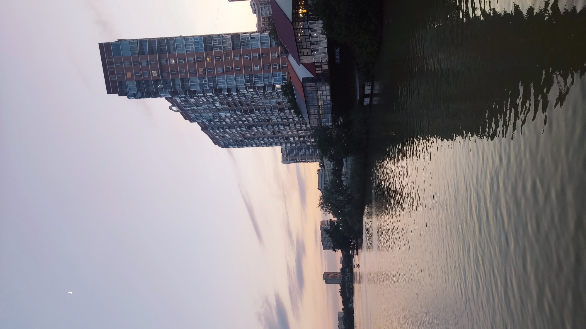 The image depicts a waterfront scene in the city of Krasnodar, Russia, likely during twilight. A large, modern residential building with a reddish-brown facade and numerous windows dominates the right side of the frame. Below this building, a row of single-story structures with glass facades and colorful roofs, possibly restaurants or cafes, is visible along the water's edge. The water in the foreground reflects the sky and the buildings. In the background, across the river, several other multi-story buildings are visible. The sky is a soft gradient of pastel colors, with a thin crescent moon visible in the upper left quadrant. A few boats are on the water, and trees line the riverbank. The overall impression is one of urban development adjacent to a natural waterway.