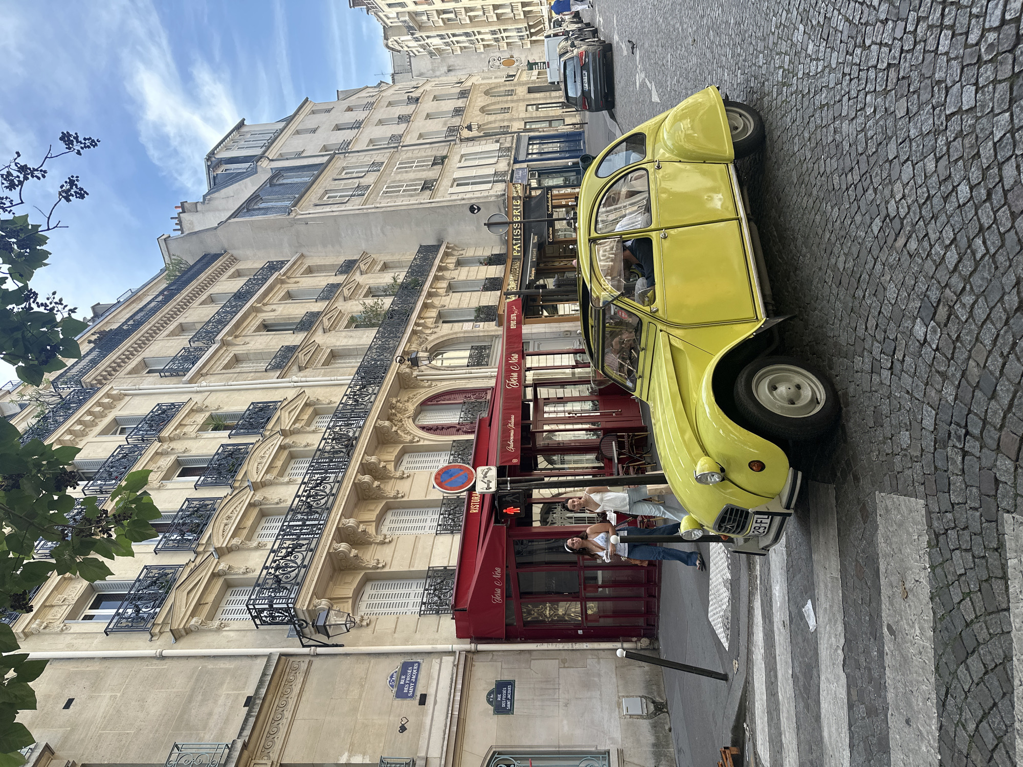This media file depicts a street scene in Paris, France, likely during the daytime. The primary focus is a bright yellow vintage Citroën 2CV car parked on a cobblestone street at a crosswalk. Two young women are standing near the car, one on the sidewalk and another in the street, appearing to interact or pose.
Behind the car, a multi-story Parisian building with traditional architecture lines the street. The building features ornate balconies with wrought-iron railings and numerous windows, some with shutters. Several storefronts are visible at the ground level, including one with a red awning and signage that reads "Terra Nera," suggesting it might be a restaurant or cafe. Another sign, "Patisserie Cuisine," indicates a pastry shop.
The sky is blue with wispy white clouds, suggesting fair weather. The street is paved with cobblestones, a characteristic feature of many European cities, and the crosswalk markings are clearly visible.
Visible text includes street signs for "RUE DES FOSSÉS SAINT JACQUES," indicating the location. Other text on storefronts is also present, such as "Terra Nera," "Gatronomia tatiana," and "Patisserie Cuisine," along with parts of other signs and the car's license plate, "29-FL". The overall atmosphere appears casual and relaxed, capturing a moment of everyday life in a Parisian setting.