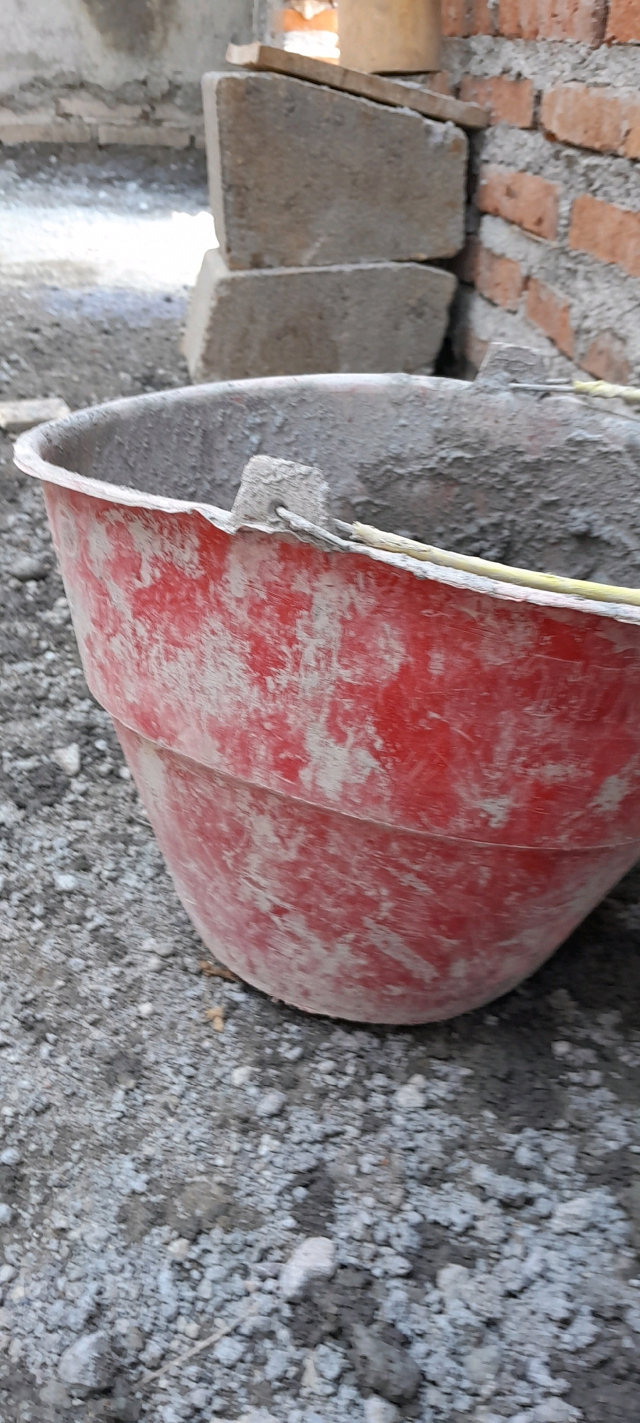 A red plastic mortar tub, partially filled with cement and debris, sits on a gravel surface in Karangduren, Indonesia. The tub's exterior is stained with dried cement. In the background, several concrete blocks are stacked, and a brick wall is under construction. The scene depicts a construction site environment.