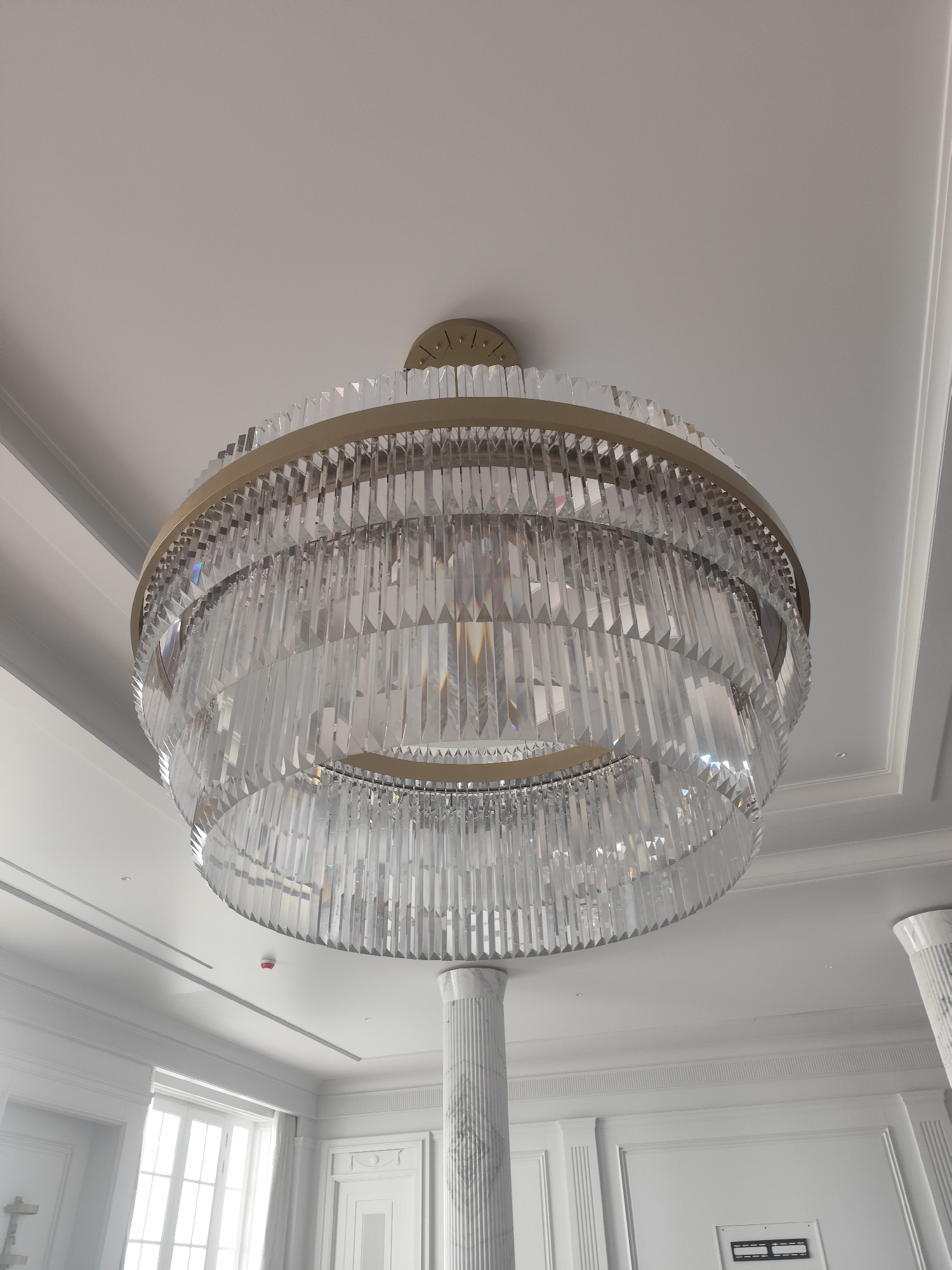 A low-angle shot shows a large, multi-tiered chandelier hanging from the ceiling of a room in an unspecified location. The chandelier features a gold-colored frame suspending numerous clear, rectangular crystal prisms. Below the chandelier are columns that extend from the floor to the ceiling, with visible architectural details. In the background, there are windows and various wall decorations, suggesting an upscale interior design. The entire setting is illuminated by a neutral light, enhancing the reflective quality of the chandelier and the white color scheme of the room.
