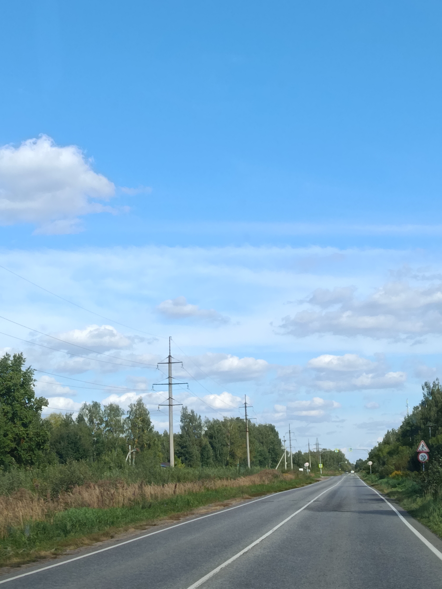 The image shows a two-lane asphalt road with white lane markings, curving slightly to the left as it recedes into the distance. The road is flanked by dense green trees and vegetation on both sides. Overhead power lines and transmission towers are visible along the left side of the road. A few traffic signs are present, including a pedestrian crossing sign with a speed limit of 70 km/h on the right side of the road. The sky is a clear blue with scattered white clouds. The scene appears to be a rural or semi-rural setting outside the city of Vereya, Russia, likely on a clear day. No people or vehicles are prominently featured in the image.