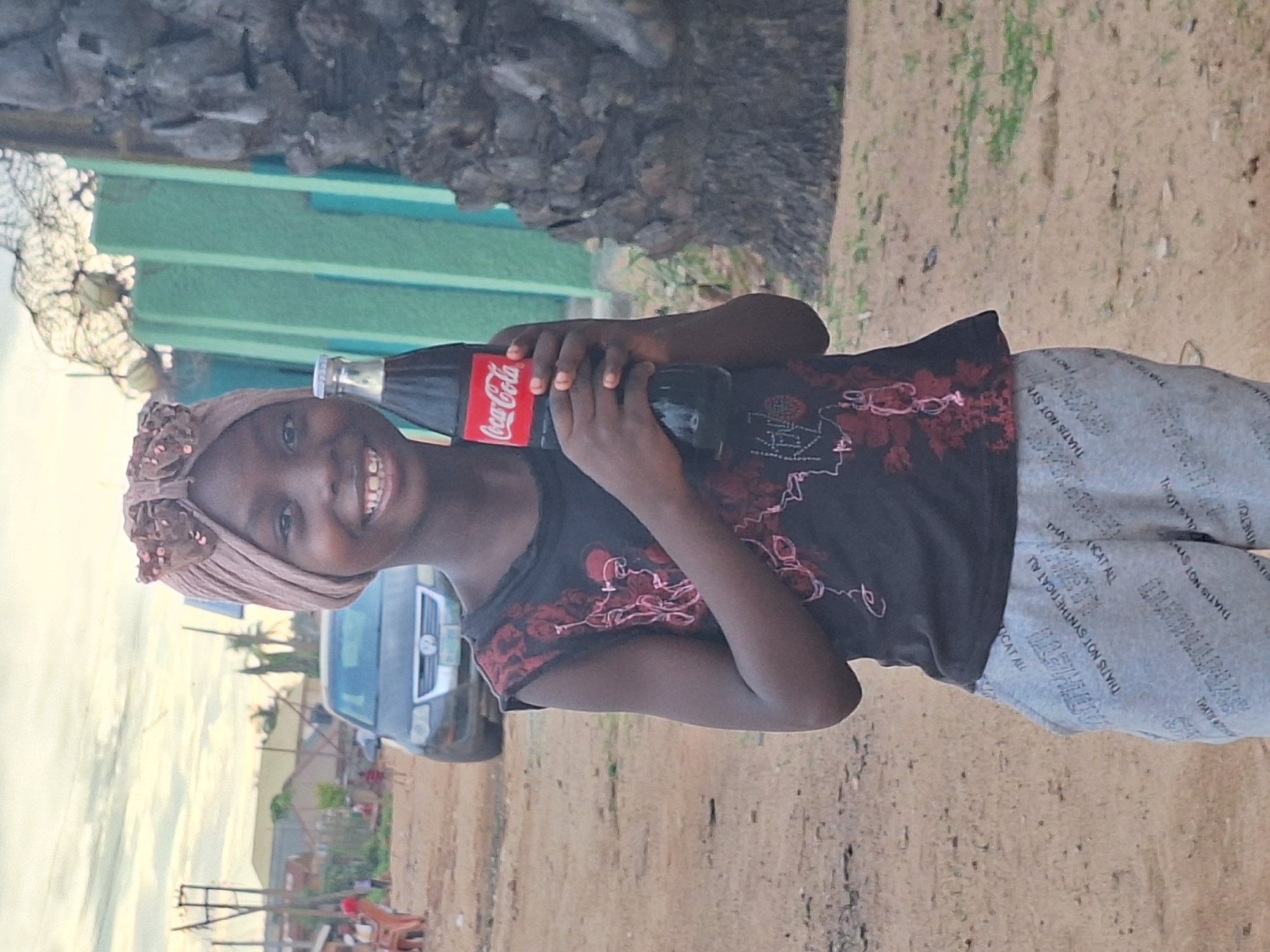 A young girl in Sabon Tasha, Nigeria, stands outside holding a large Coca-Cola bottle on her shoulder. She wears a brown head wrap, a dark t-shirt with a floral design, and grey pants printed with "THATIS NOT SYNTHETICAL AT ALL." The background shows a dirt ground, a green building, a vehicle, and other buildings in the distance under a cloudy sky.