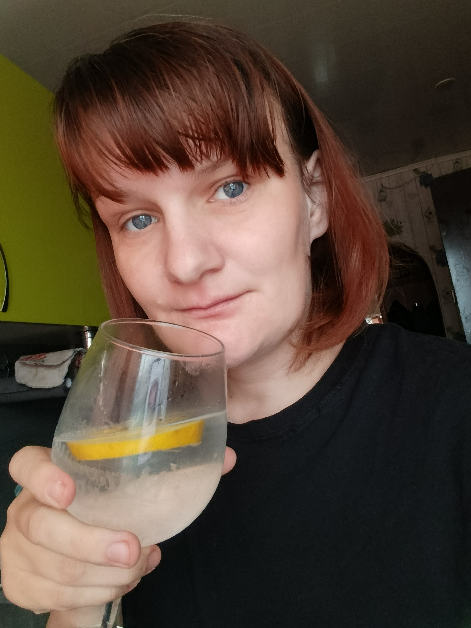 A person with reddish-brown hair and blue eyes holds a glass of water with a lemon slice. The person is wearing a black t-shirt. The background includes a green wall, part of a kitchen counter, and a wall with light-colored wallpaper. The location is Фоки, Russia.