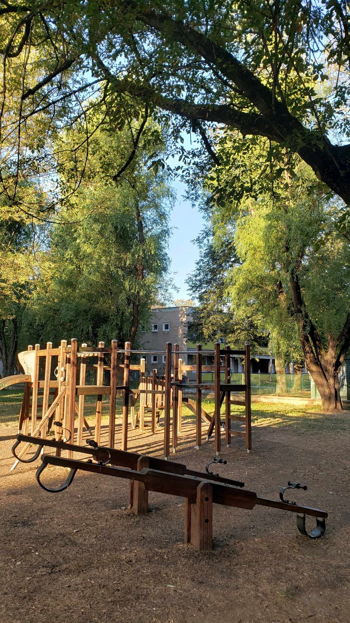 The image depicts a playground scene on a sunny day, likely in Prešov, Slovakia, given the context. In the foreground, a wooden seesaw with two seats is prominently featured. Behind the seesaw, a wooden playground structure, possibly a climbing frame or fort, stands with various platforms and railings. The ground is covered with dirt and small stones, and patches of grass are visible in the background. A building with a light-colored facade and dark windows can be seen behind the playground equipment. Mature trees with lush green foliage frame the scene, casting dappled shadows on the ground. The overall impression is one of a well-maintained outdoor play area on a bright, clear day.