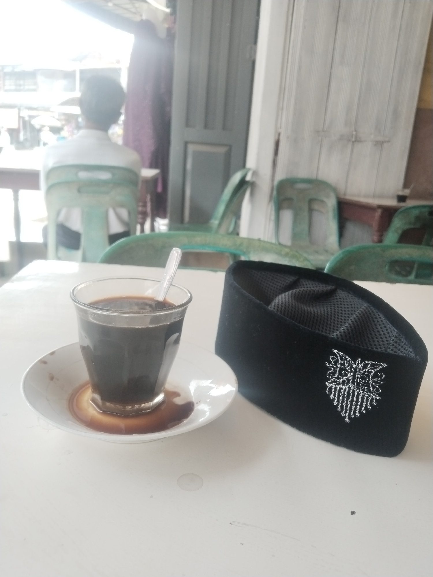 The image captures a casual scene at what appears to be an outdoor or semi-outdoor cafe or warung in Keudegeudong, Indonesia, under bright daylight conditions.
In the foreground, resting on a white table, is a clear glass of black coffee with a spoon in it, placed on a white saucer. There are visible coffee spills or drips on the saucer. To the right of the coffee, a black peci or songkok hat is placed. The hat has an intricate white embroidered emblem or crest on its front, featuring stylized, symmetrical patterns, and a mesh lining is visible inside.
The background is softly out of focus, depicting the bustling environment of a local establishment. A person, seen from the back, is seated on a green plastic chair, facing away from the camera. Several other green plastic chairs are scattered around, suggesting multiple seating arrangements. Behind the person and chairs, elements of a building, possibly a wooden door or wall, are visible. The bright lighting suggests it is daytime, likely a sunny day. The overall atmosphere conveys a relaxed, everyday moment at a local gathering spot.