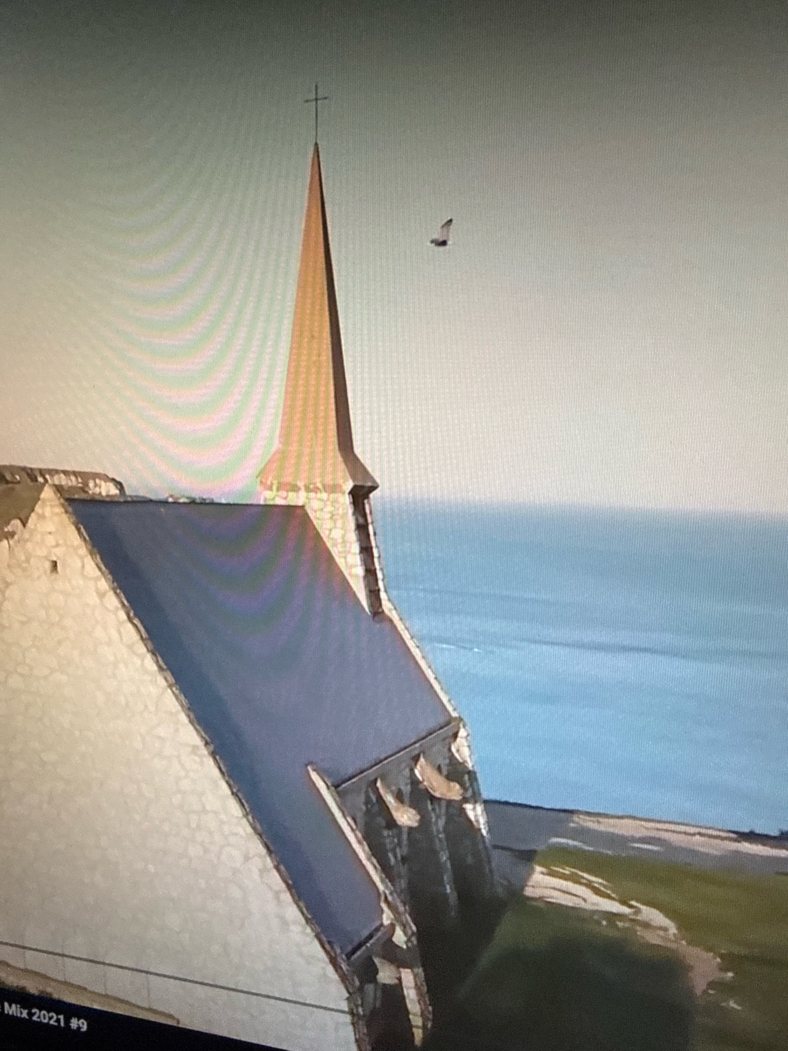 An aerial, high-angle shot shows a church steeple and roof against a bright sky and a body of water. The steeple is tall and conical, topped with a cross. A bird is flying in the sky near the steeple. The church is made of stone with a dark roof. In the foreground, there are green and brown patches of land. The bottom left corner of the image shows the text "Mix 2021 #9". The scene appears to be a coastal area, possibly in Netanya, Israel, given the visual characteristics of the building and surrounding landscape often seen in this region.