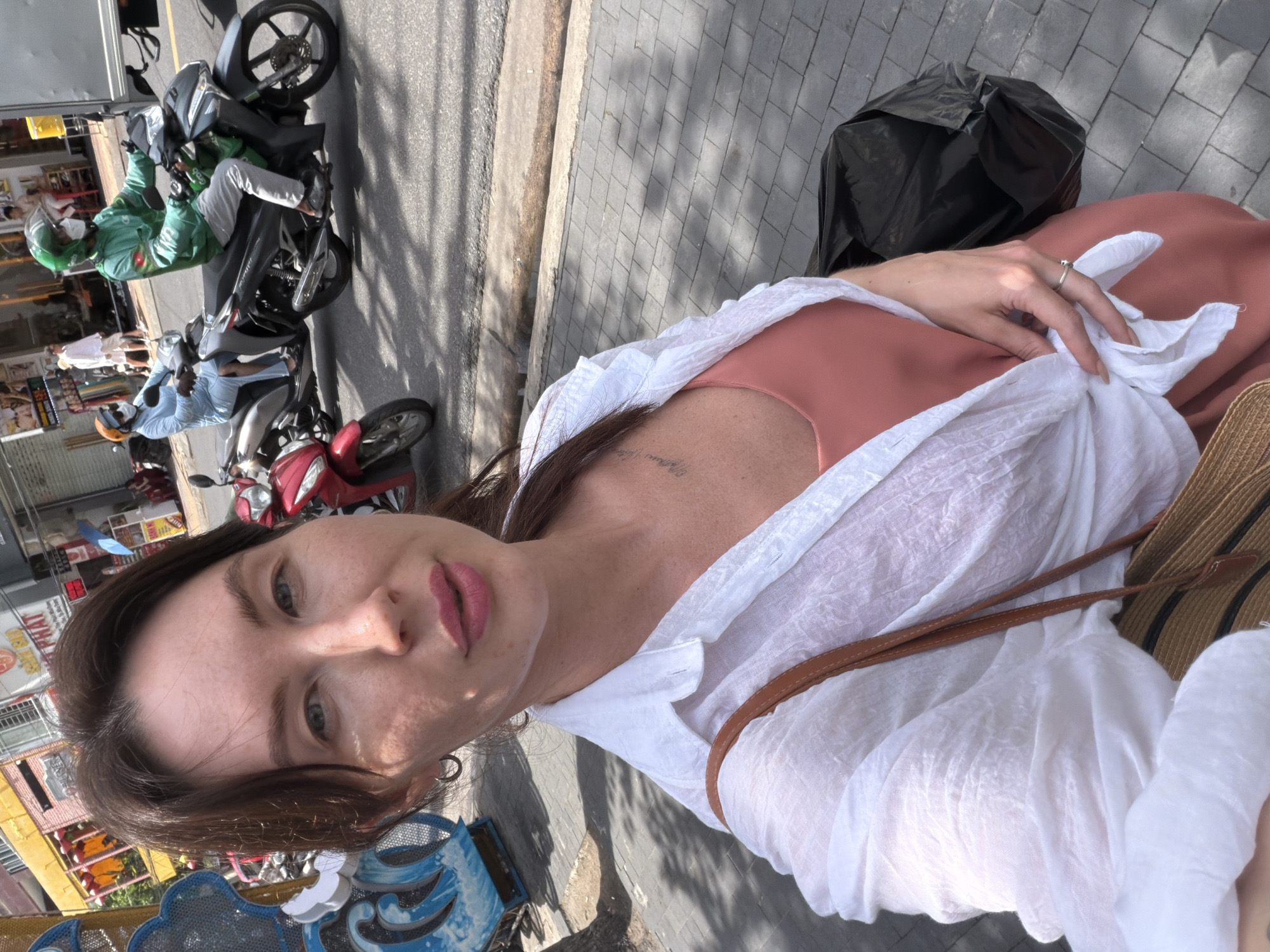 A woman with brown hair takes a selfie while standing on a sidewalk next to a street. She is wearing a white open button-down shirt tied at the waist over a coral pink tank top, a brown woven bag with leather straps over her shoulder, and a silver ring on her right hand. In the background, two men on motorcycles are visible. The man on the left is wearing a light blue outfit and a white mask. The man on the right is wearing a green jacket, grey pants, a green helmet, and a white mask, and is riding a dark colored motorcycle. Behind the motorcycles, there are buildings with awnings, signs, and a blue and white decorative structure on the left. The ground is paved with cobblestones. The scene appears to be in Nha Trang Ward, Vietnam, based on the architecture and street activity.