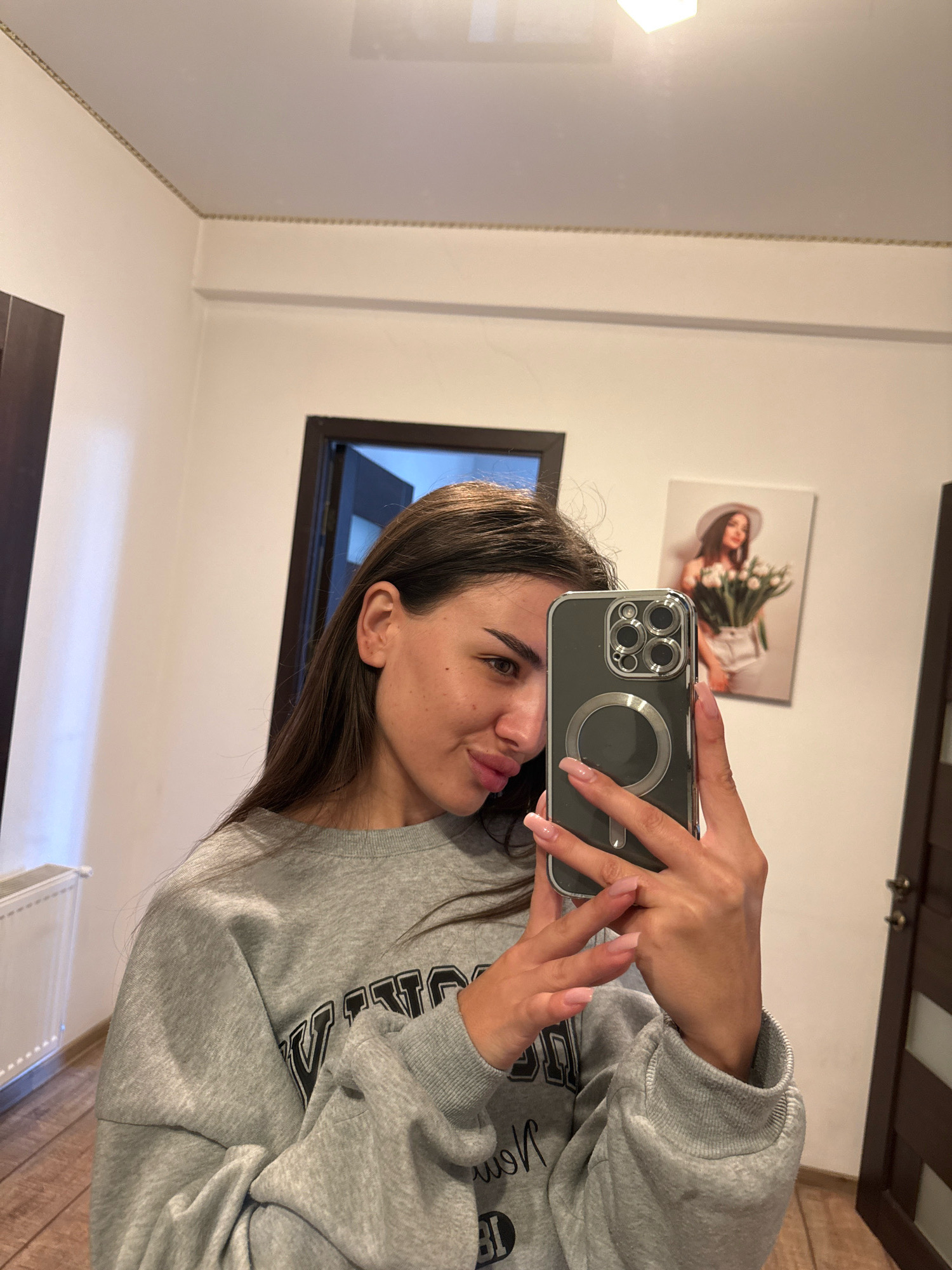 A young woman with long brown hair takes a mirror selfie indoors. She is wearing a grey sweatshirt and is holding a smartphone in her right hand. The phone has a grey case with multiple camera lenses and a circular magnetic ring. Her left hand is positioned near the phone. In the background, there is a framed picture of a woman wearing a white hat and holding a bouquet of white flowers. There are also doors and a radiator visible in the room. The lighting suggests an indoor setting, possibly a home or hotel room. There is no indication of the location being in Chișinău, Moldova.