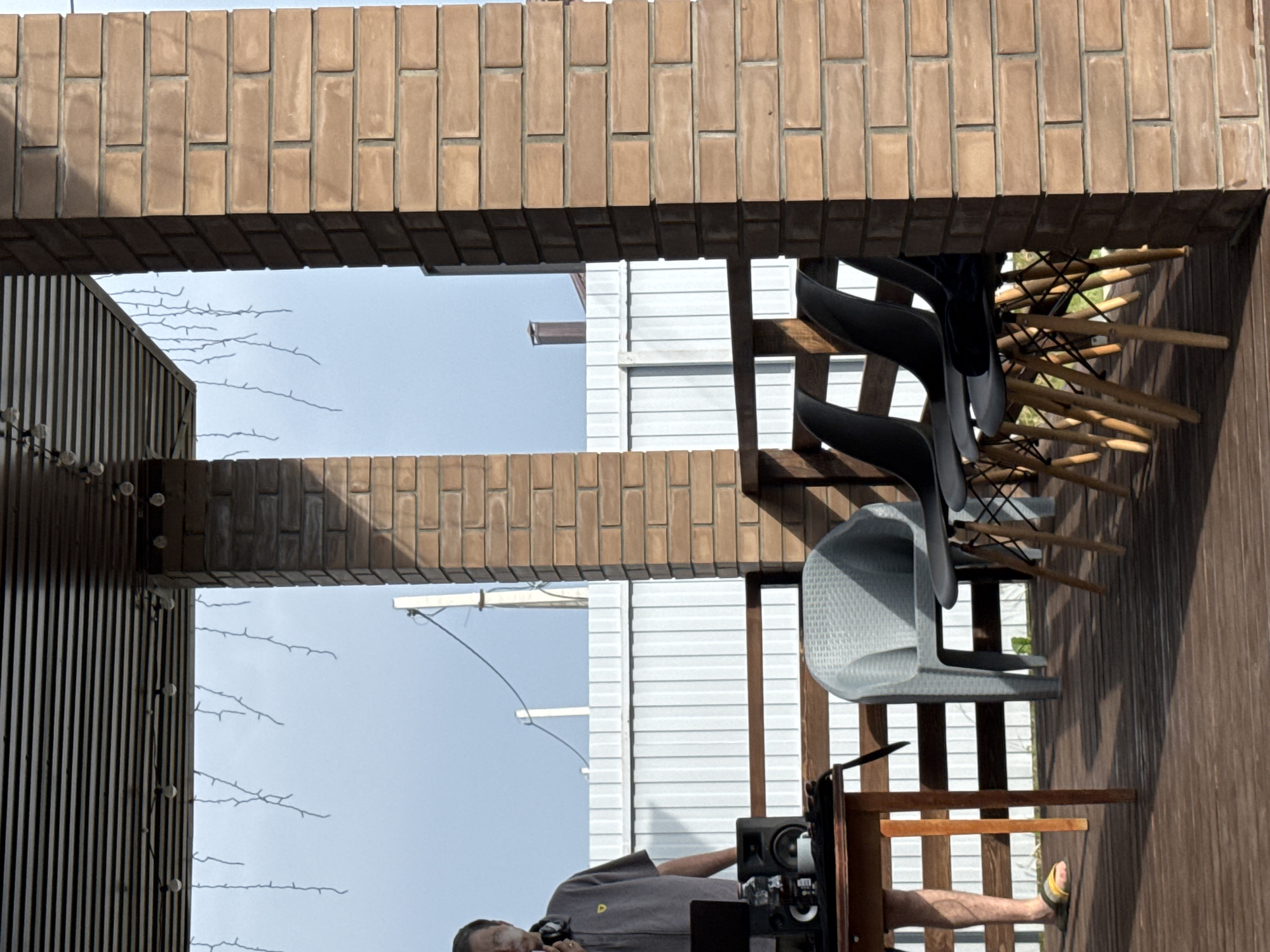 A man, wearing a gray collared shirt and yellow sandals, stands on a wooden deck in front of a table with a laptop and speakers. He is holding a cigarette and wearing headphones around his neck. Several chairs are arranged on the deck. The deck is attached to a brick building. The background consists of a white fence and a cloudy sky. The location is in the city of Dyomino, Russia.