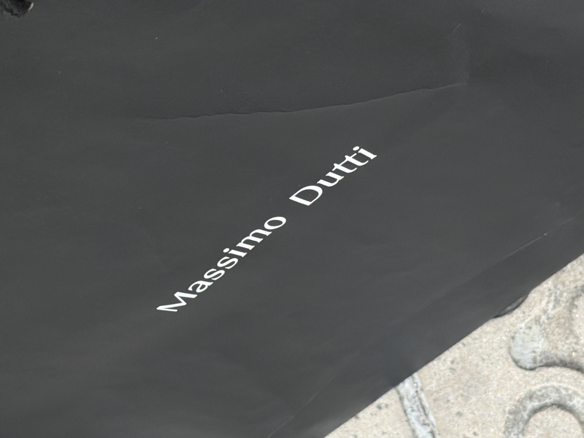 The image shows a black paper shopping bag with the white logo "Massimo Dutti" printed diagonally across it. The bag is resting on a light-colored paved surface that appears to be a sidewalk or street, with some circular indentations visible. The background is dark and out of focus. The scene appears to be outdoors in an urban setting, possibly in Barcelona, Spain, given the context. There are no people or visible actions in the image.