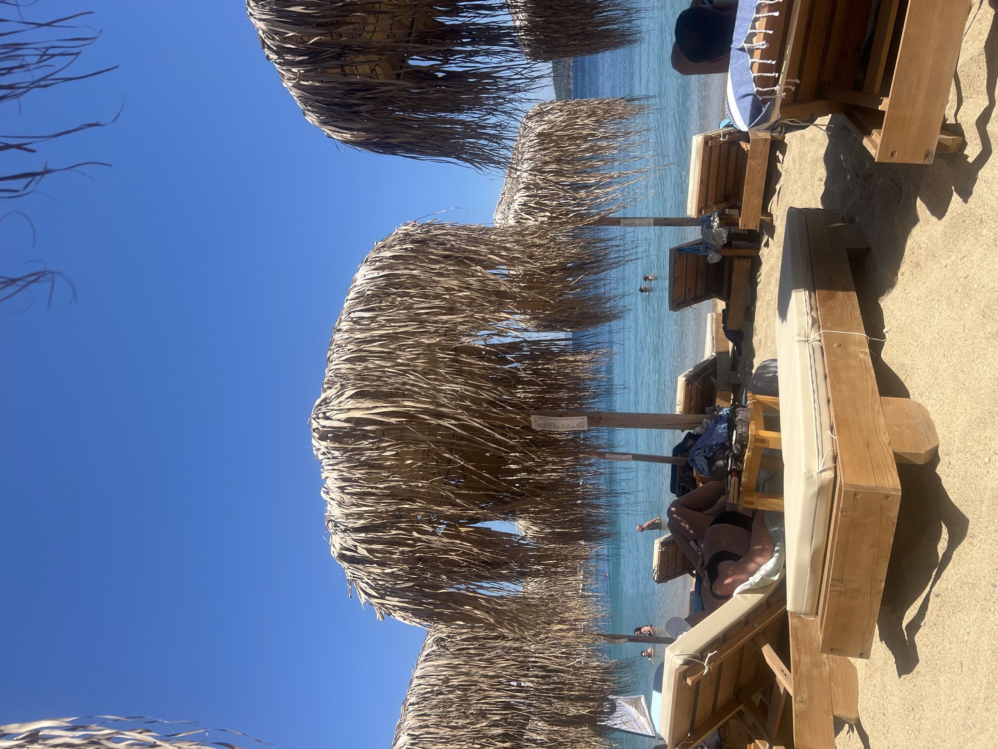 The image depicts a beach scene in Achladias, Greece, under a clear blue sky. Several thatched umbrellas provide shade over wooden lounge chairs arranged on the sand. In the background, the calm, turquoise sea is visible, with a few people swimming and wading. Some individuals are relaxing on the lounge chairs, while others are partially obscured by the umbrellas. The overall atmosphere is one of leisure and relaxation on a sunny day.