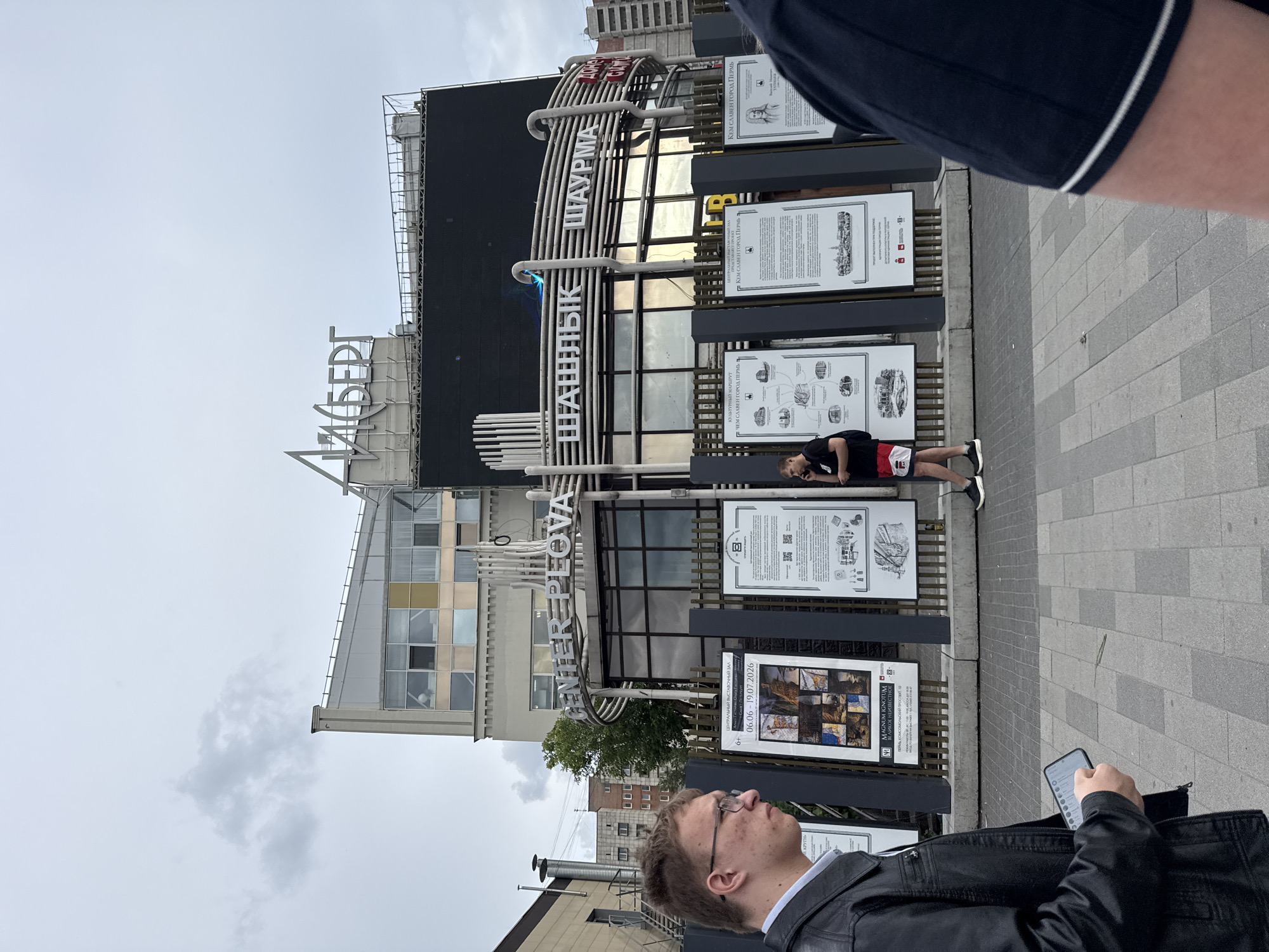 This media file depicts an outdoor scene in Perm, Russia, featuring a building with prominent signage and several display boards. The building's facade is modern, with large windows and a distinctive metal structure adorned with white lettering. One sign clearly reads "ШАШЛЫК ШАУРМА," indicating a food establishment, likely a shawarma and shashlik place. Another sign spells out "АЖІБЕРГ."
Several vertical display boards are arranged in front of the building. These boards appear to be informational or promotional, featuring text and images. One board is partially obscured but shows a portrait, while others display text related to "КЕМ СЛАВЕН ГОРОД ПЕРМЬ" (Whom is the city of Perm famous for) and a date range of "06.06-19.07.2026." Another board shows a title "MAGNUM AGNUM IGNOTUM" and a collage of images.
In the foreground, a young person wearing a black t-shirt and red and white shorts is visible, looking down. To the right, the back of another person wearing a dark blue shirt is seen, with their arm extended holding a smartphone. A third person, a young man wearing glasses and a black leather jacket, is in the lower right portion of the frame, also looking upwards and holding a smartphone.
The weather appears to be overcast, with grey clouds filling the sky. The ground is paved with grey tiles. The overall impression is of a public space in a city, possibly a cultural or commercial area, with people interacting with the displays and the environment. The time of day is not explicitly clear but suggests daytime.