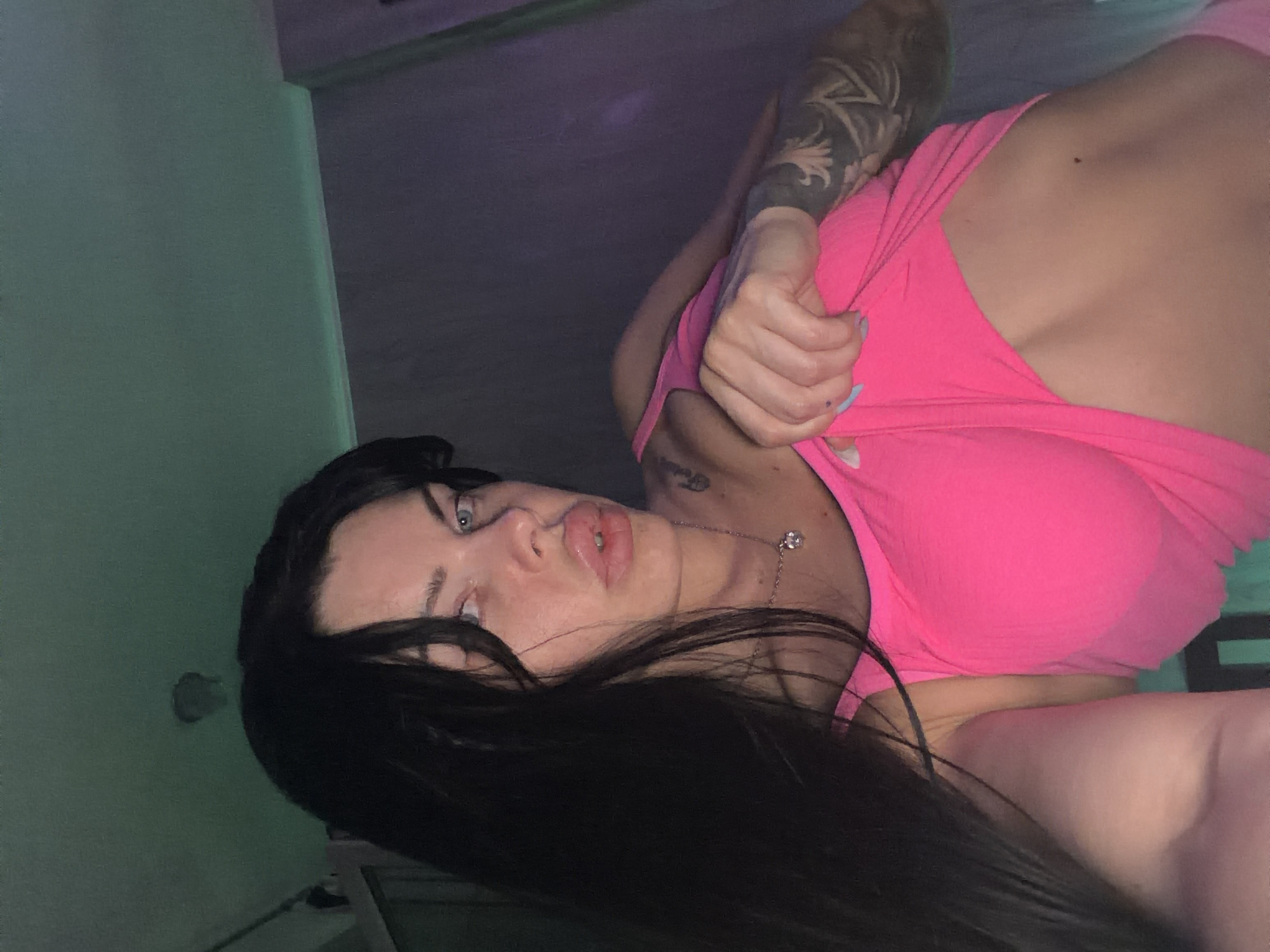 The media file is a low-angle, close-up, selfie-style photo of a young woman with long, dark hair and blue eyes. She is wearing a bright pink top and appears to be lying down, looking up at the camera. Her lips are slightly parted, and her gaze is directed towards the viewer. She has a tattoo on her upper left arm and a delicate necklace with a charm around her neck. The lighting is dim and atmospheric, with hints of purple and green. The background is indistinct, suggesting an indoor setting. There is no visible text in the image. The overall mood is intimate and suggestive.