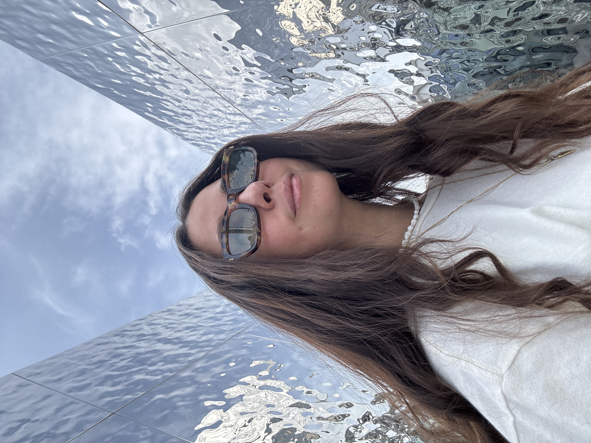 A young woman is centered in the frame with dark, wavy, shoulder-length hair, wearing sunglasses and a white sweater. The photograph is taken at a tilted, upward angle. She appears to be standing between two mirrored walls that create an illusion of flowing water. The sky, with light clouds, is reflected in the mirrored surfaces. The location is Kazan, Russia, indicated by the distinctive architecture and mirrored art installation. The person is looking slightly up towards the camera, suggesting she is taking a selfie.