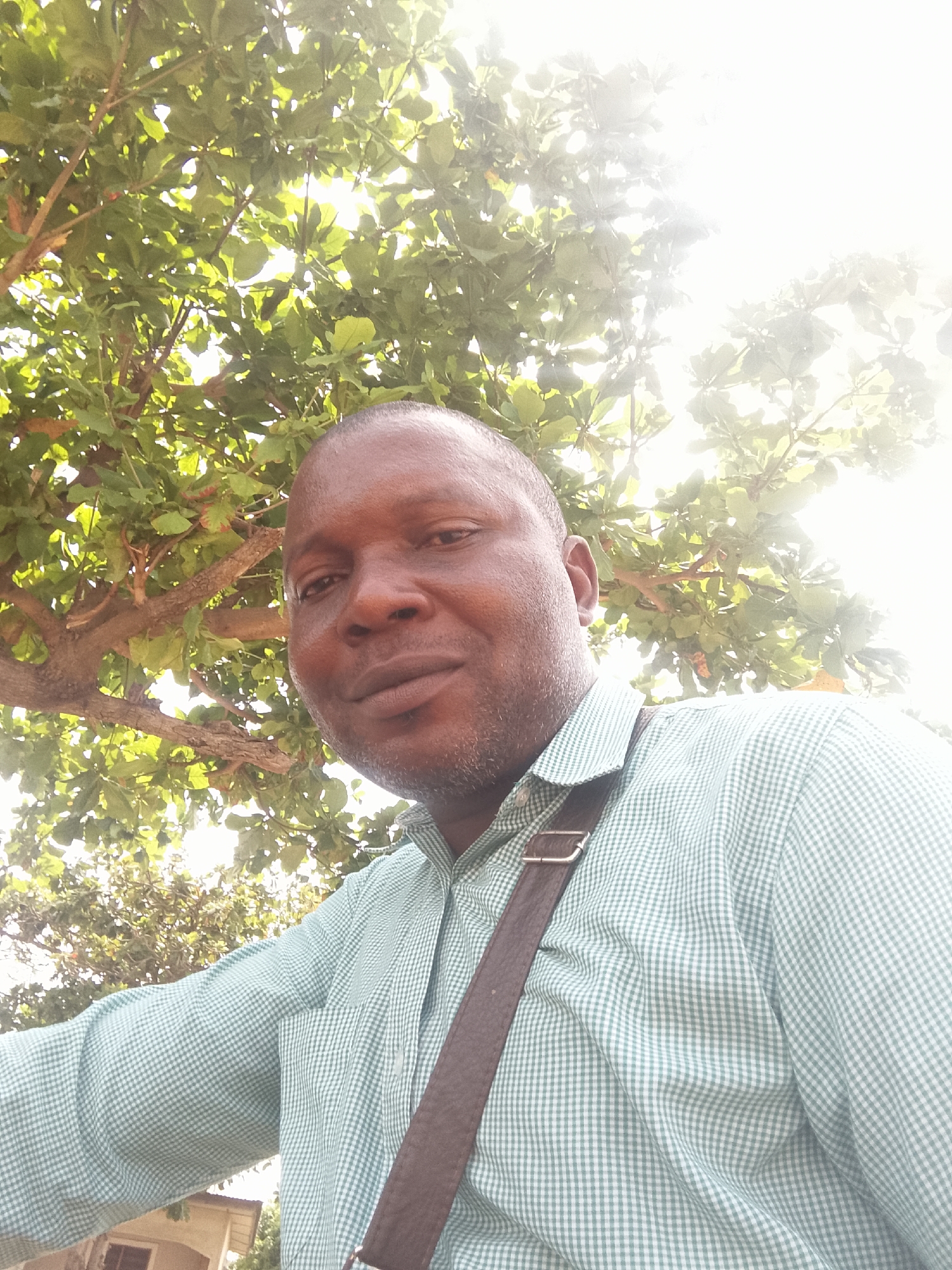 A man is shown from the chest up, wearing a light green and white checkered long-sleeved shirt and a brown strap over his shoulder. He has dark skin, a shaved head, and a slight beard. He is standing under a tree with dense green leaves and branches that fill the upper portion of the frame. The sunlight is bright, causing glare in the upper right. In the background is a building. The location is Unknown, Unknown.