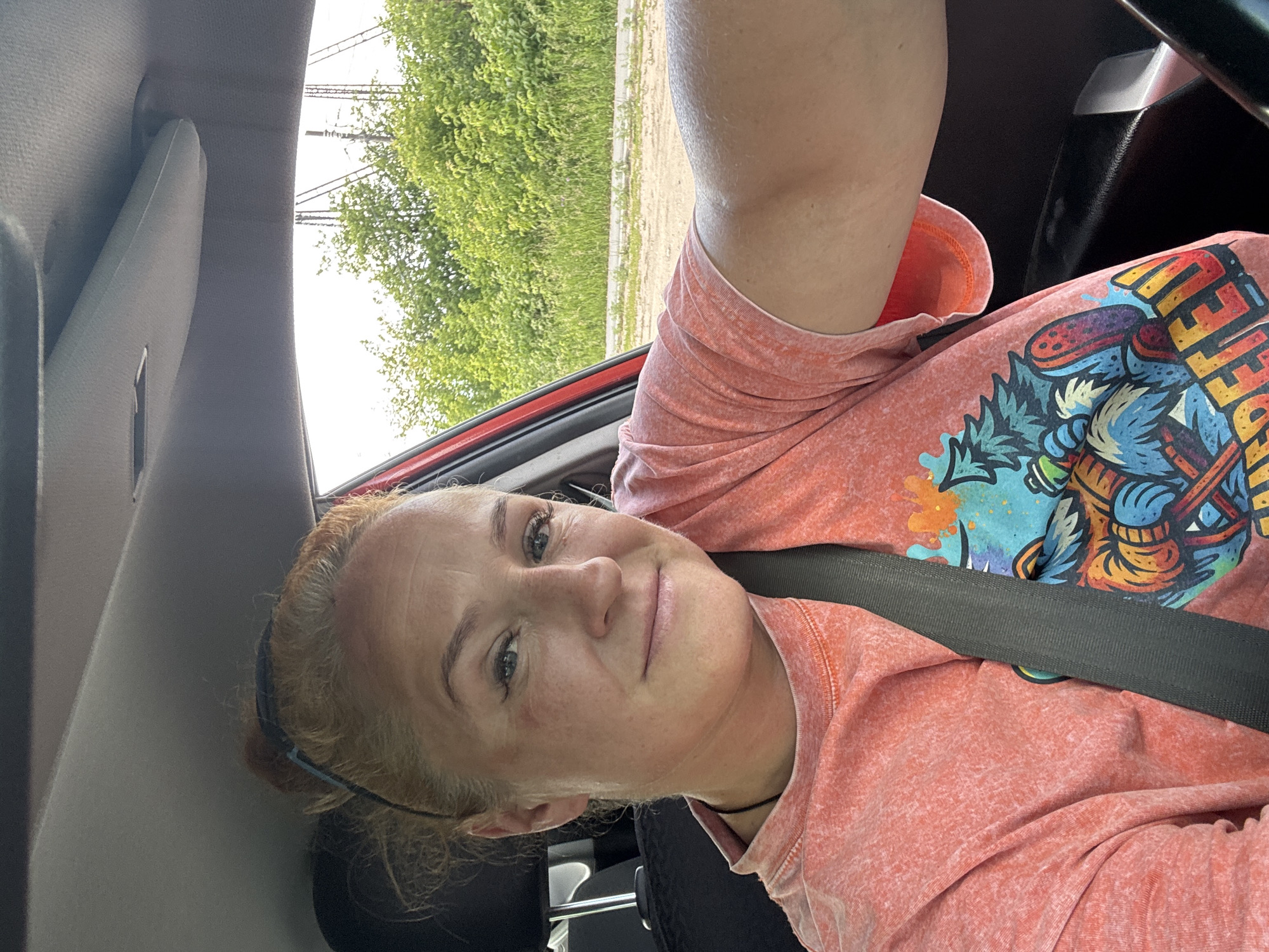 An outdoor, slightly overexposed, front-facing selfie taken from inside a red car shows a Caucasian woman with blonde hair pulled back in a bun, wearing a coral-colored t-shirt with a graphic print, and a seatbelt. The woman's facial features are visible, including her eyes, nose, and mouth. She is looking towards the camera with a slight smile. The background of the image shows lush green trees and vegetation, a dirt road, and utility poles against a bright sky. The interior of the car, including the car ceiling, a portion of the steering wheel, and a car seat, is also visible. The image is likely taken in the city of Chaikovsky, Russia, given the context provided.