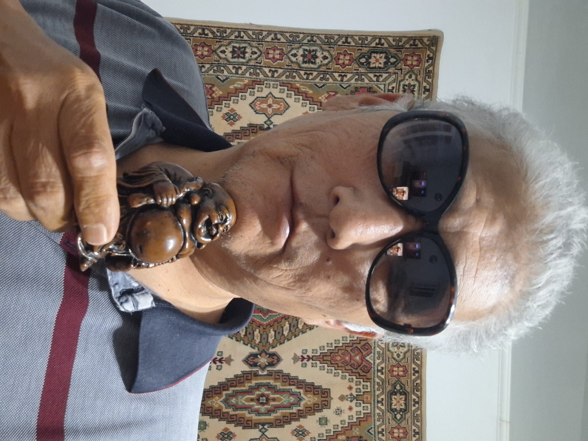 The image shows a close-up, front-facing selfie of a person wearing large, dark-rimmed sunglasses and a gray collared shirt with a burgundy stripe. The person has gray hair and is holding a small, wooden figurine of a laughing Buddha. The background features a tapestry with a geometric pattern. The lighting appears to be indoor. The provided information states that the image is located in Almaty, Kazakhstan.