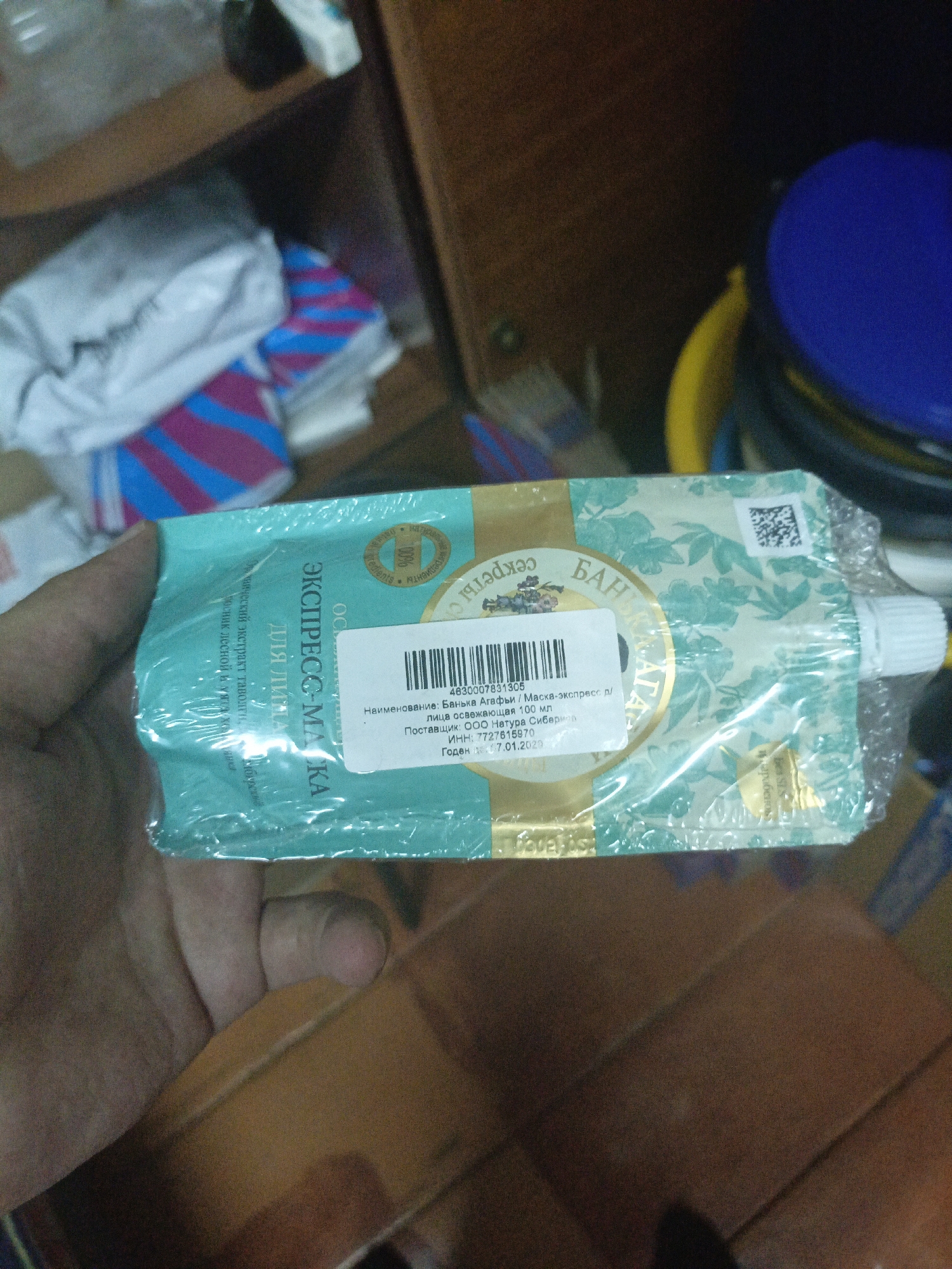 A person's hand holds a tube of cosmetic product, likely a face mask, that is still sealed in plastic packaging. The tube is a light teal color with a pattern of green leaves and flowers. The text on the tube is in Russian and appears to be a product name and description. A white label with a barcode and more Russian text is affixed to the front of the tube. The background is blurred but appears to be an indoor setting with wooden shelves and some other objects, including a blue plastic container. The product is labeled as "ЭКСПРЕСС-МАСКА" (Express Mask) and mentions "Баня Агафьи" (Banya Agafya), a Russian cosmetic brand. The label also includes production information, including the manufacturer "ООО Натура Сиберика" (LLC Natura Siberica) and a production date of 17.01.2020. The image does not contain information to determine its location in the city of Ухманы, Russia.