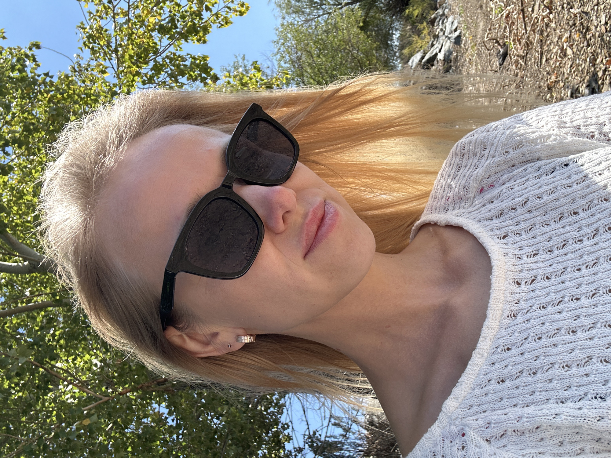 A young woman with blonde hair and sunglasses is pictured outdoors. She is wearing a white knit top. The background consists of trees, foliage, and a rocky, arid terrain. The scene appears to be located in the city of Gornyi Erik, Russia, based on the provided context.