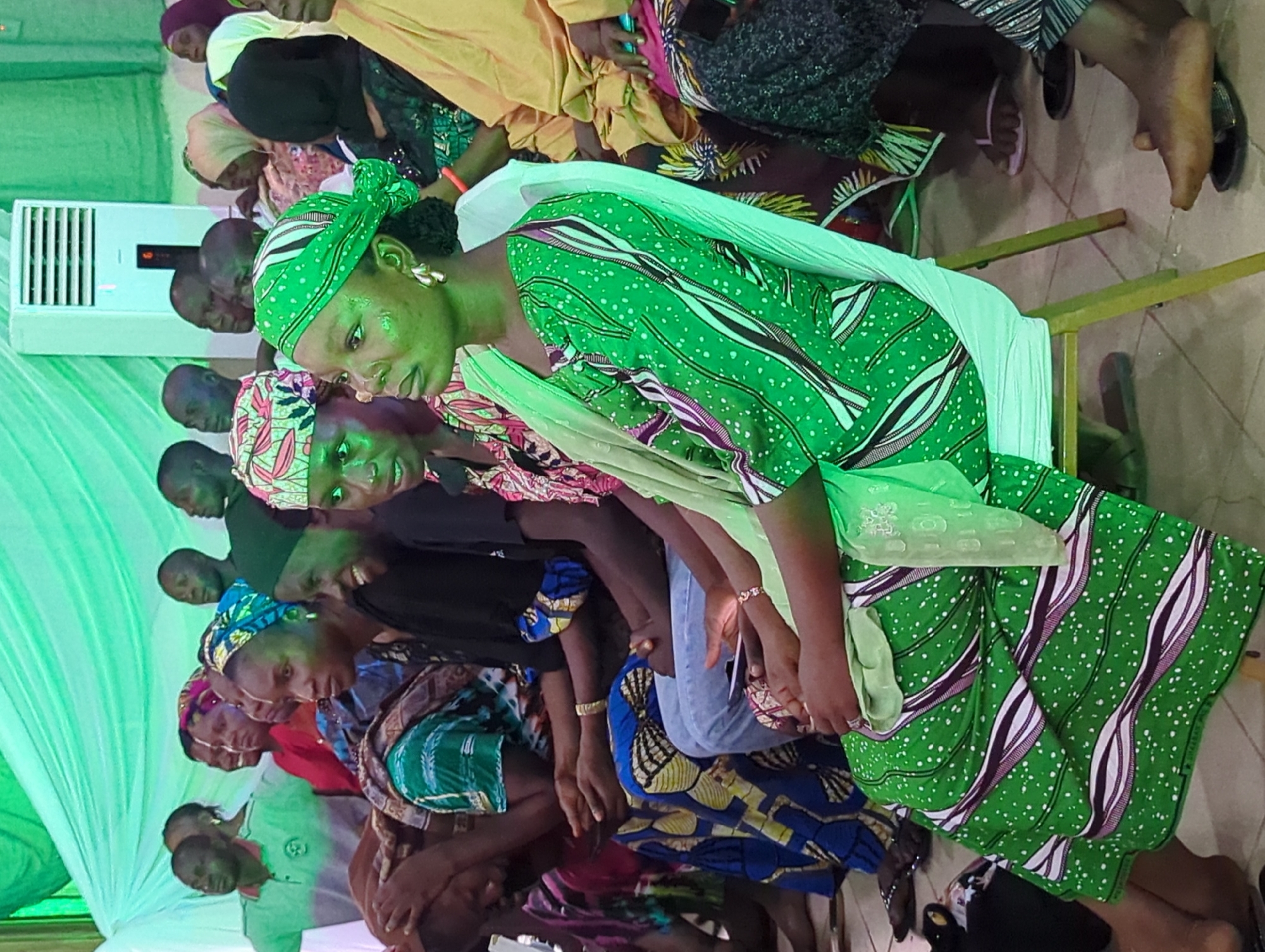 A group of people are seated indoors in Unknown, Unknown. The primary subject is a woman wearing a green dress with white and purple accents and a matching head wrap. The surrounding individuals are similarly dressed in colorful garments. An air conditioner is visible in the background, and the setting appears to be a gathering or event.