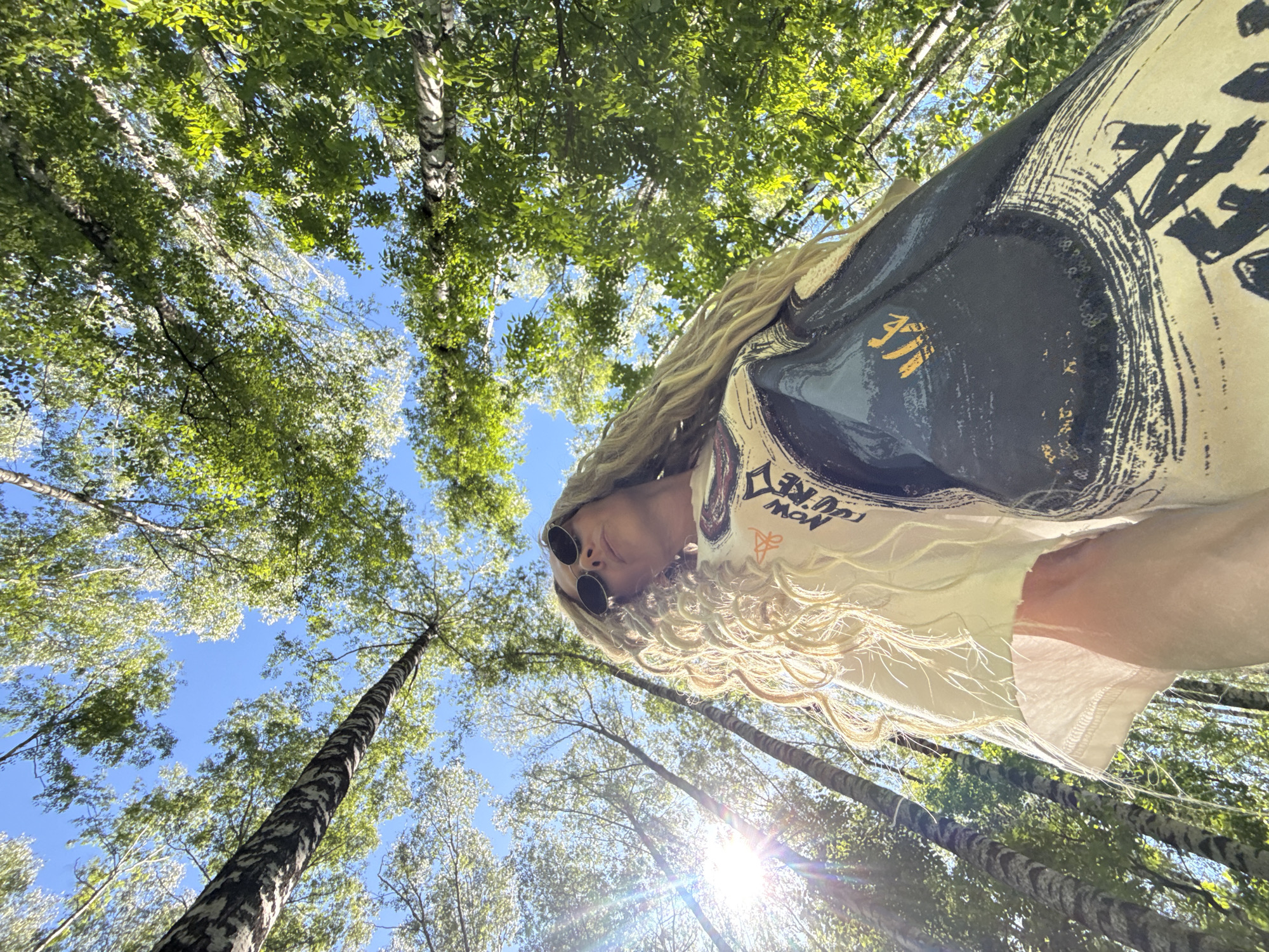 The image depicts a female subject photographed from a low-angle perspective within a sunlit forest, identified as being in the city of Заволжье, Russia. The subject, positioned in the lower-center and extending diagonally upwards to the right, has long, voluminous, light blonde textured hair and wears dark, round sunglasses. She is dressed in a light-colored (possibly off-white) t-shirt with a visibly frayed or ripped left sleeve hem. Over or with the t-shirt, she wears a dark-colored garment, likely a dress or long skirt, which features large, light-colored abstract brushstroke patterns, circular motifs with sequins, and partial text (including "NOW CURE" visible on the t-shirt, and "CAL" and a stylized yellow/gold symbol on the dark garment). Sunlight emanates from the upper-left of the frame, creating a pronounced sunburst and lens flare, illuminating the subject and the trees. The background consists of a dense canopy of tall, slender trees, identifiable as birches by their distinct bark patterns, with abundant green foliage against a clear blue sky. Multiple tree trunks extend vertically throughout the frame, emphasizing the upward perspective.