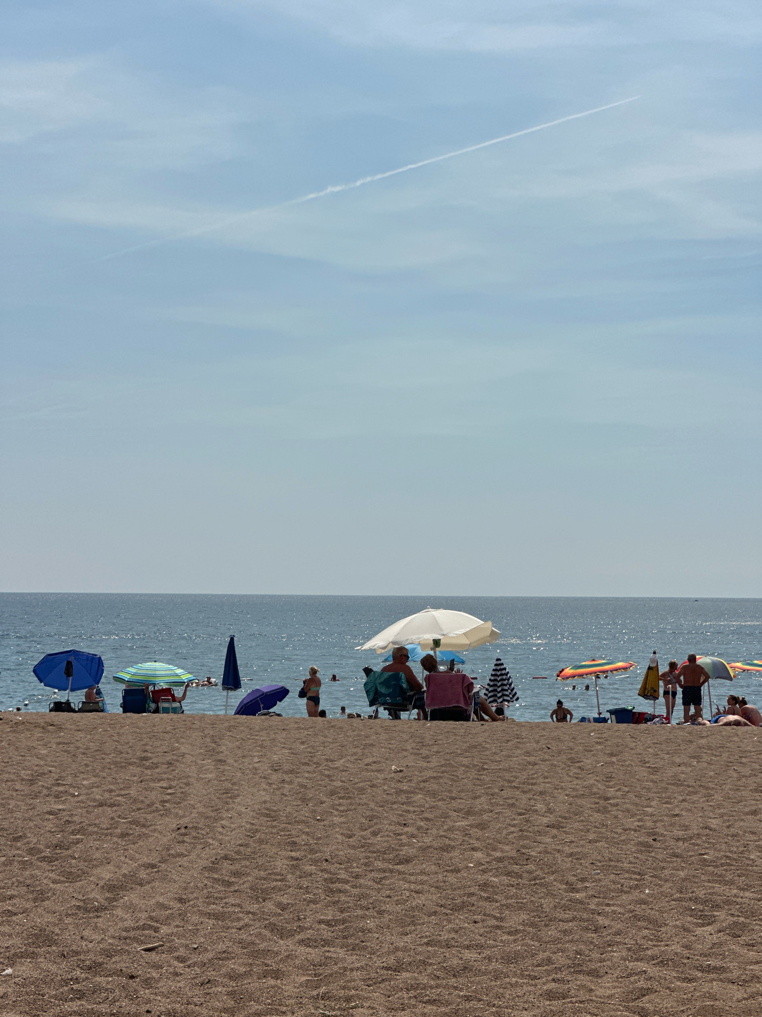 The image depicts a crowded beach on a sunny day. The foreground is dominated by a sandy beach, with some footprints and debris visible. In the mid-ground, a line of beach umbrellas shades groups of people. Several individuals are seated in beach chairs, while others stand or walk along the shoreline. The ocean stretches out in the background, calm and blue, with small waves lapping at the shore. A thin contrail is visible in the bright blue sky. The scene appears to be a popular tourist destination, with a lively atmosphere. Based on the context provided, this scene is located in Boreti, Montenegro.