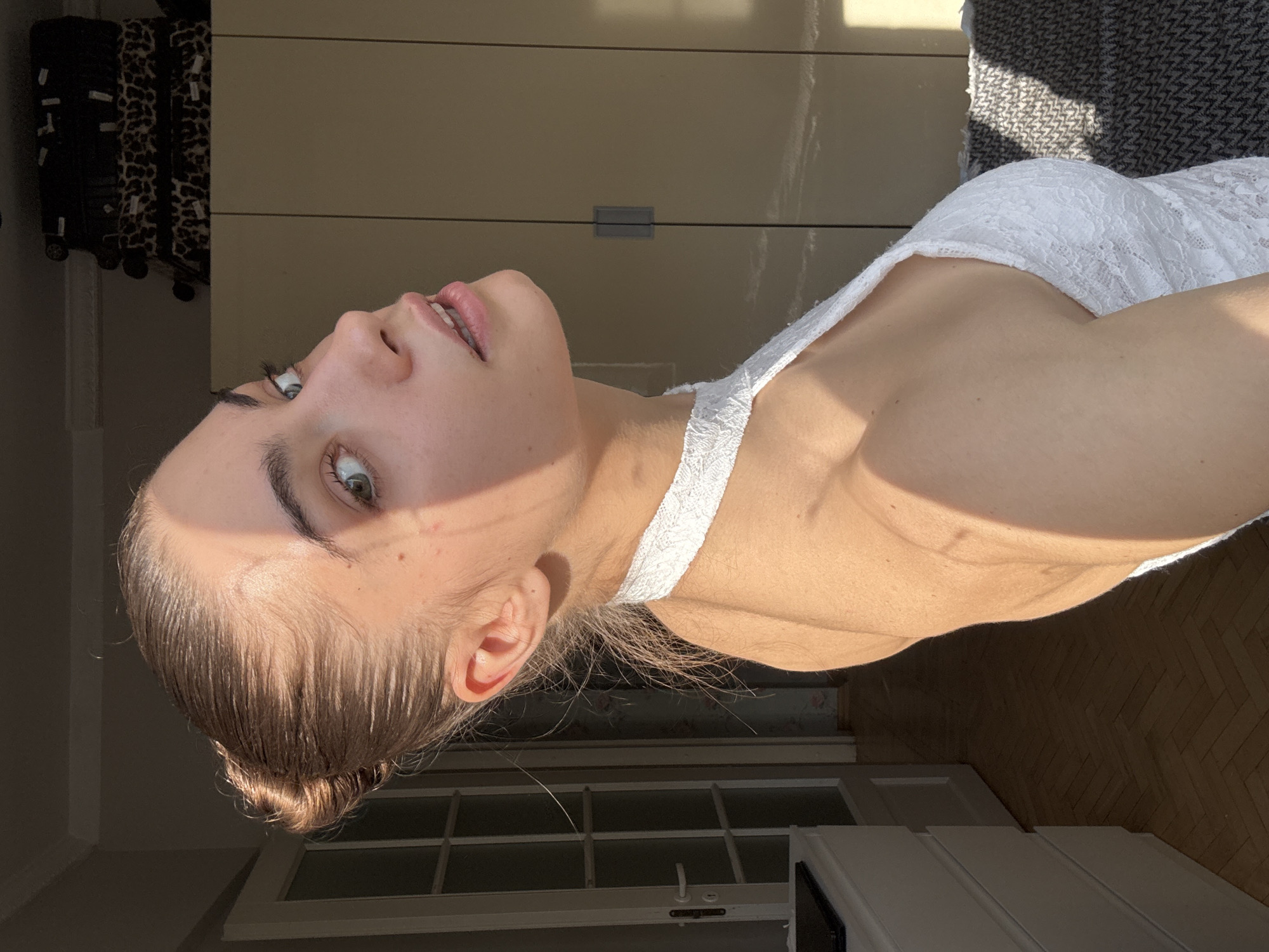 A young woman with fair skin, light brown hair pulled back in a bun, and green eyes is positioned in a room. She is wearing a white lace top. The background includes a closet with a leopard print suitcase, a window, and hardwood flooring. The location is not identifiable from the image as Moscow, Russia.