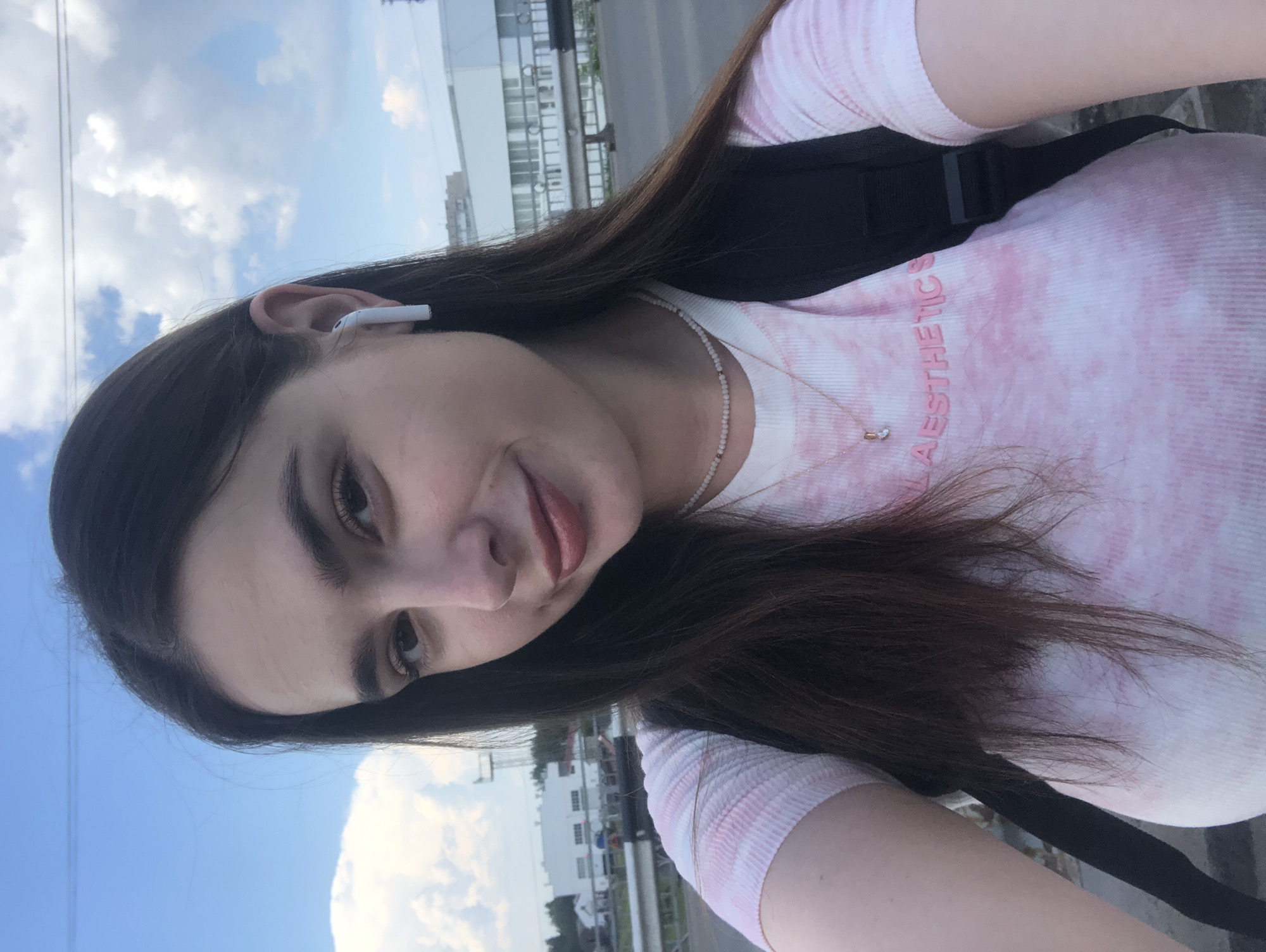 A young woman with long, dark brown hair is pictured in a close-up, slightly upward-angled selfie. She is wearing a pink and white tie-dye ribbed t-shirt with the word "AESTHETICS" embroidered in pink on the front, a delicate pearl necklace, and a small gold necklace with a clear pendant. She has on white wireless earbuds and is wearing a black backpack, visible over her left shoulder. The background suggests an outdoor, urban or industrial setting with a cloudy blue sky, a bridge or elevated walkway, and a large white building with a row of windows. The presence of power lines and industrial structures indicates a location within the city of Chaikovsky, Russia. The lighting appears to be natural daylight.