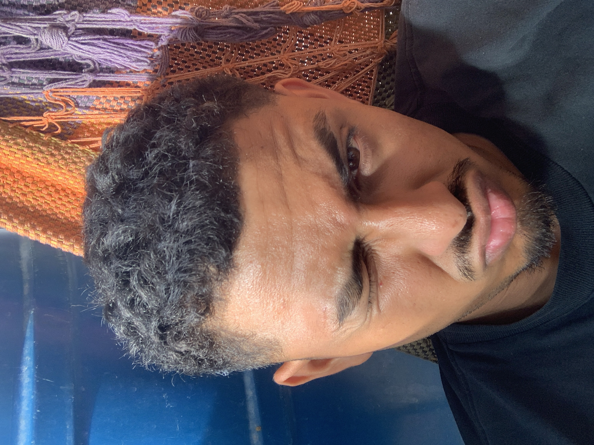 A medium close-up selfie shows a young man of color with dark curly hair and a goatee winking and pouting. He is wearing a black t-shirt. The background is blurred, with a blue object on the left and a macrame wall hanging with orange and purple tassels on the right. The lighting is bright and appears to be natural. The man's skin is reflective, indicating he may be sweating. He is positioned in what is described as a location in Santa Ana, Venezuela.