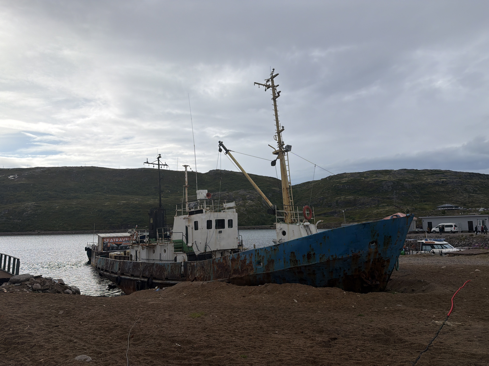A rusted, blue and white ship is beached on a sandy shore in Teriberka, Russia, with a body of water to its left and a hilly, sparsely vegetated landscape to its right. The ship, appearing derelict, has a tall mast with various antennas and equipment. To the left of the ship, a small dock made of wood and rocks is visible. In the background, several buildings and vehicles are present, including a white van and what appears to be a small passenger boat. The sky is overcast with gray clouds. The overall scene depicts a quiet, possibly industrial or maritime, setting in a coastal town.