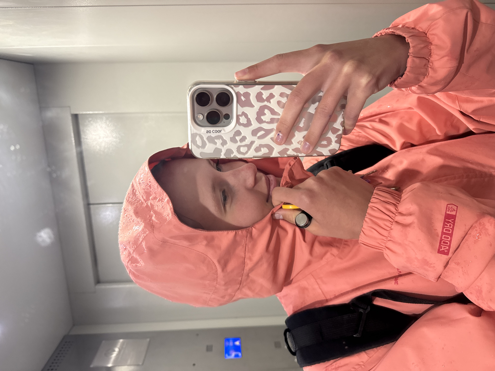 A person in a pink hooded jacket is taking a selfie in what appears to be an elevator. The phone has a white case with a light brown leopard print design. The person is wearing a backpack with black straps. Droplets of water are visible on the jacket. The elevator interior is simple, with metal walls. There is no information available in the image to confirm the specific location in Chaikovsky, Russia.