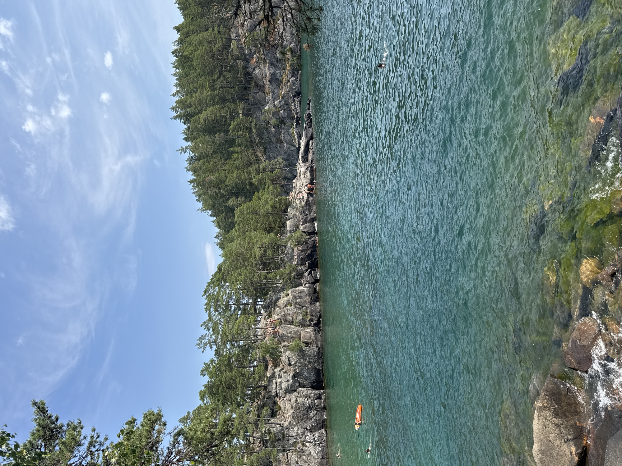 This is a landscape photograph taken on a clear, sunny day, likely during the summer months, at a rocky shoreline in or near Karlsborg, Sweden. The image captures a body of water with a rocky, tree-lined cliff face in the background.
The foreground features a rocky shore with clear, turquoise water revealing submerged rocks and vegetation. The water is slightly choppy, creating small ripples on the surface.
In the middle ground, the water is populated with several individuals engaged in recreational activities. Some are swimming, while at least one person is on an orange inflatable raft. A person with a snorkel and mask is visible swimming further out.
The background consists of a steep, rugged cliff face covered in dense green coniferous trees. There are several people visible on the rocks at the top of the cliff, some of whom appear to be standing at the edge, possibly preparing to jump into the water. A few more individuals are seated or standing on the rocky ledges.
The sky above is blue with scattered white, wispy clouds, indicating fair weather. The overall impression is a scene of outdoor leisure and activity in a natural, scenic environment.