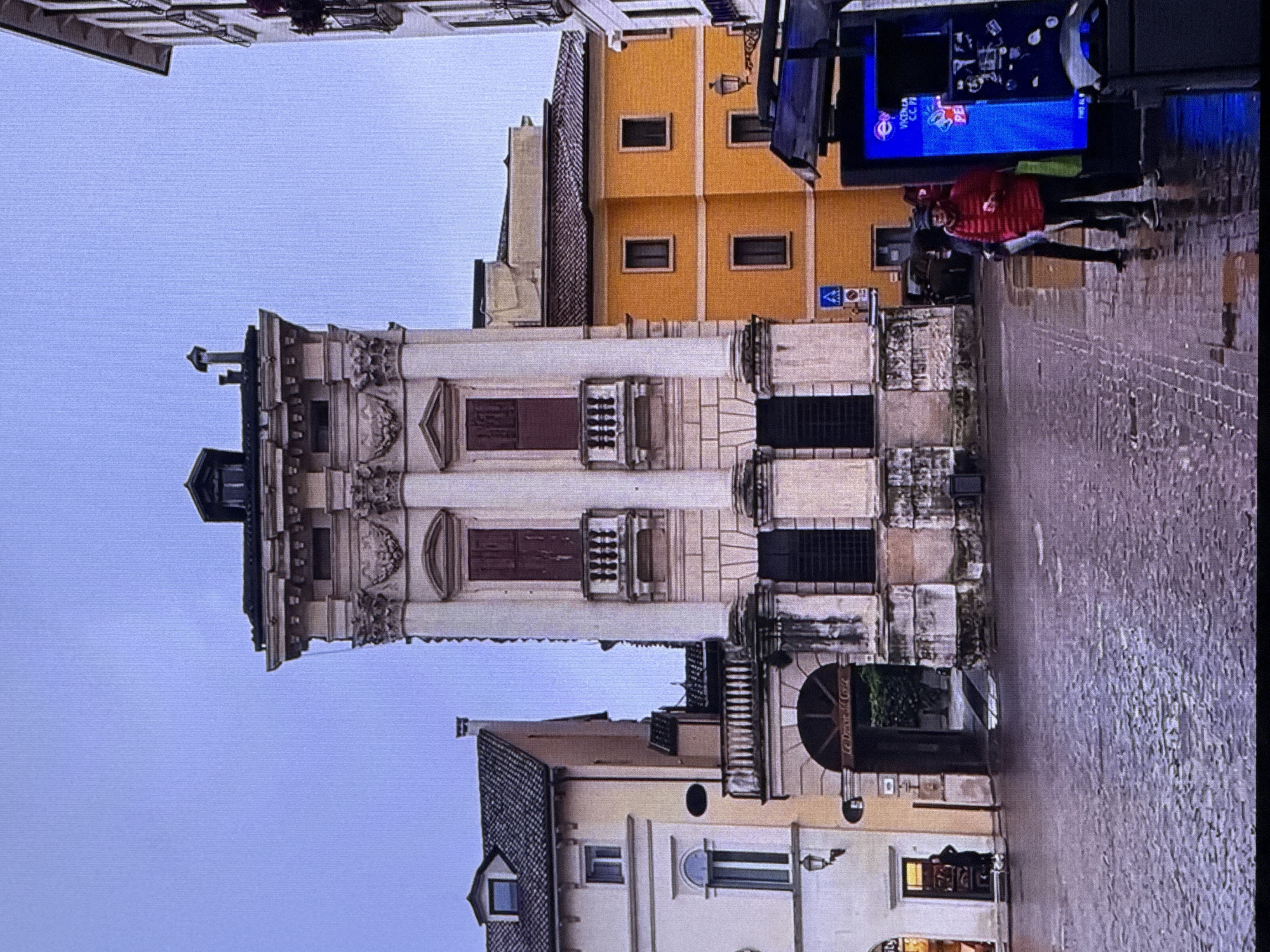 The image shows a tall, ornate building with classical architectural elements, including columns and decorative carvings. The building is beige and appears to be quite old. To the left is a lighter-colored building with a dark tiled roof and windows. To the right is a bright orange building with dark-framed windows. In the foreground, the ground is paved with cobblestones and appears wet, suggesting it has recently rained or is raining. There is a bus stop shelter with a digital advertisement screen. Several people are visible: one person in a red puffer jacket and a dark hat is standing near the bus stop, holding a red object and a green bag. Another person in dark clothing is standing behind them. In the archway of the beige building, a person is visible standing near a doorway. The sky is overcast and a pale blue. The scene is urban, with a sense of historical architecture contrasted with modern elements like the bus stop. There is no indication that this location is in Dubai, United Arab Emirates.