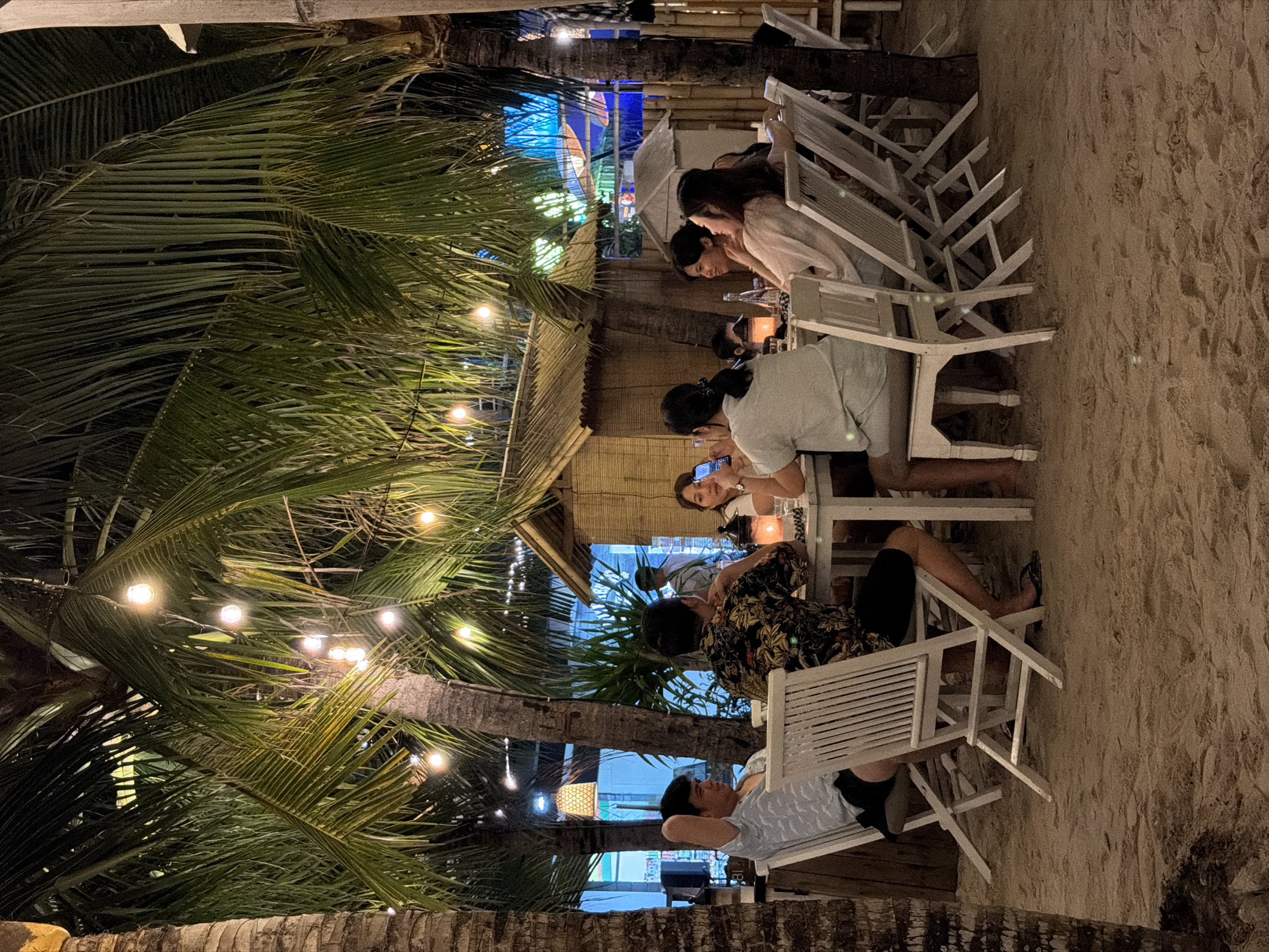 In Unknown, Indonesia, a group of six individuals is seated around a table on a sandy beach, likely at a restaurant. The setting is illuminated by strings of warm-toned lights draped through palm trees, creating an intimate evening atmosphere. A small, thatched-roof structure is visible behind the group, suggesting a tropical beachfront establishment. The diners are casually dressed, some engaged in conversation, while one person is looking at their phone. White chairs surround a white table laden with a few items, possibly drinks or tableware. The sand, illuminated by the ambient lighting, covers the ground, and palm tree trunks are prominent in the foreground and background. The overall scene depicts a relaxed dining experience in a tropical environment.
