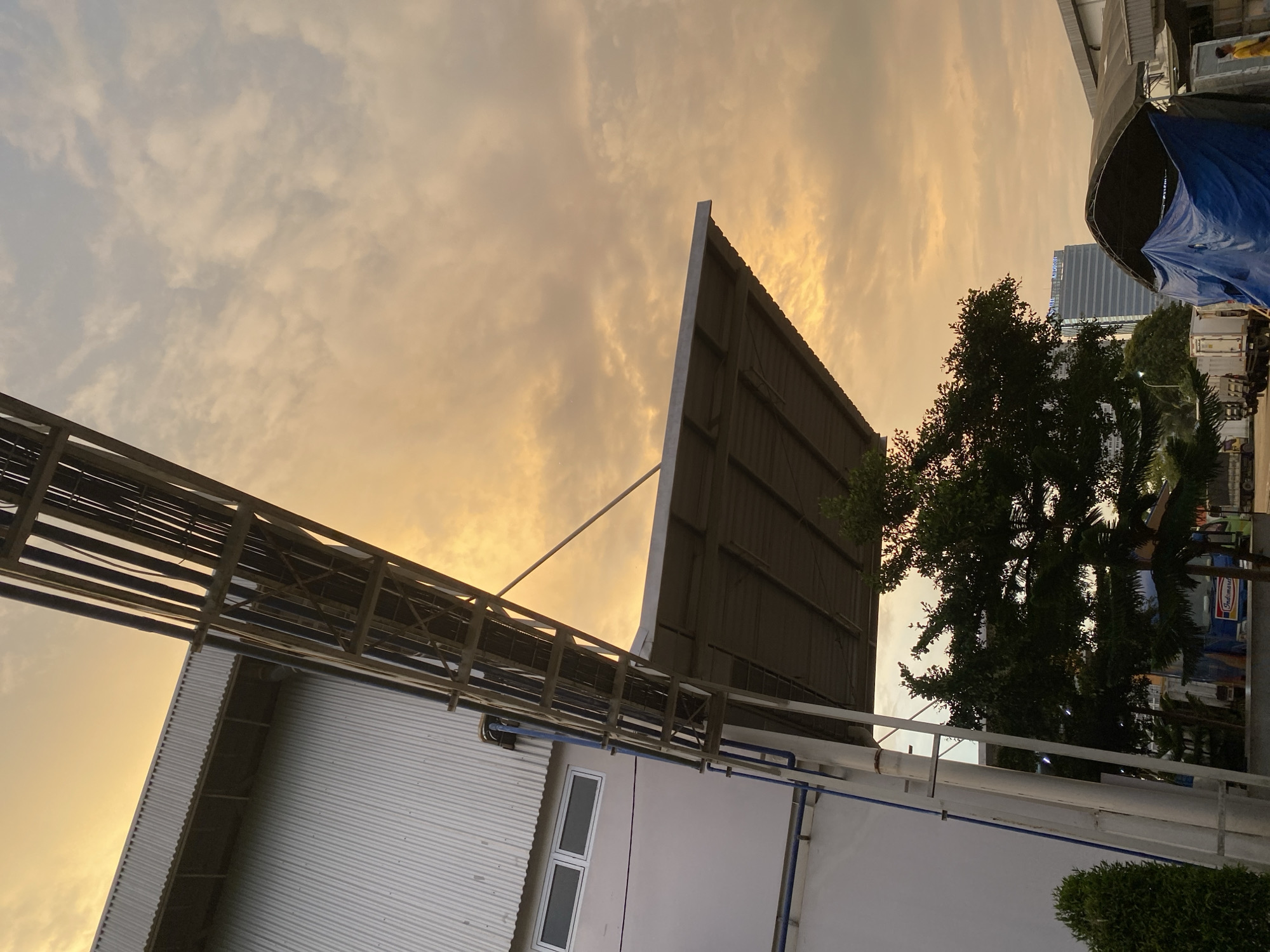 This media file shows an outdoor industrial or commercial scene at sunset. The sky is filled with warm-toned clouds, indicating either dawn or dusk. In the foreground, a corrugated metal building with a white facade is visible, with a blue pipe running along its side. Above the building, a large, dark structure, possibly a billboard or a roof, is supported by metal beams and a diagonal brace. To the right, a dense green tree stands tall, and further in the background, a modern high-rise building with reflective windows is visible. A blue tarp covers part of an object or structure in the middle ground, and several white shipping containers are parked nearby. Some faint text is visible on a sign behind the tree, which appears to say "Indomaret", a convenience store chain common in Indonesia. The overall impression is one of a developing or active industrial area bathed in the soft light of a setting sun.
