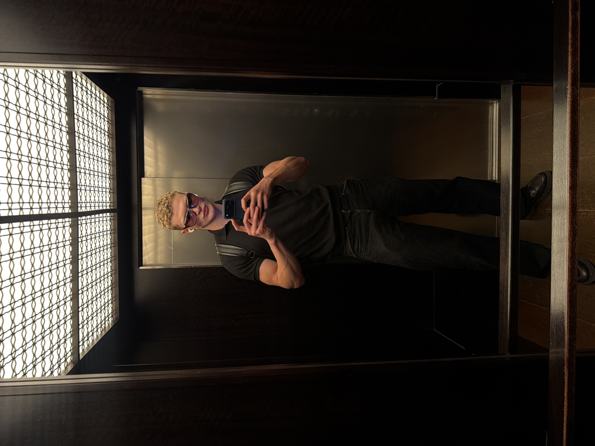 A young man with blonde, curly hair and sunglasses is taking a mirror selfie in an elevator. He is wearing a black polo shirt, black pants, and black shoes. He has a black backpack on his shoulders and is holding a smartphone in his right hand. The elevator has dark wood paneling and a metallic door. The ceiling has a decorative light fixture. The floor of the elevator is a wooden-like material, and there are some wooden ledges in the foreground. The overall impression is modern and stylish. There is no indication of a specific location in London, United Kingdom.