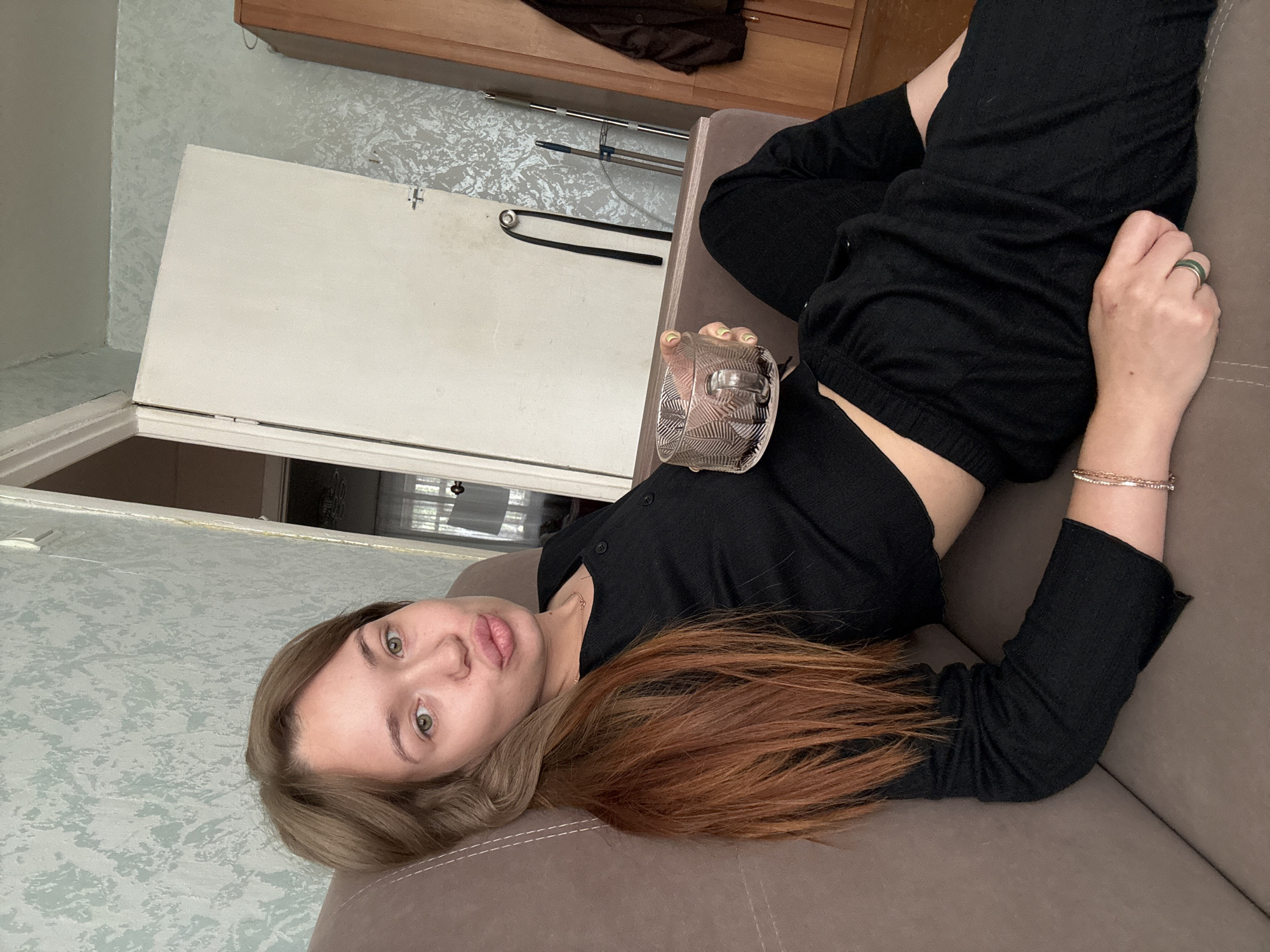 A young woman with reddish-brown hair is lying on a brown couch in a room, holding a glass of dark liquid. She is wearing a black crop top and black pants. Her lips are pursed in a pout. The room appears to be an apartment, with wallpaper on the walls, a white door, and a wooden wardrobe. The general decor suggests a home environment, possibly in Chaikovsky, Russia, given the context of the request, though the image itself does not contain explicit markers of its location. The lighting is natural, coming from a window or overhead source, casting soft shadows.