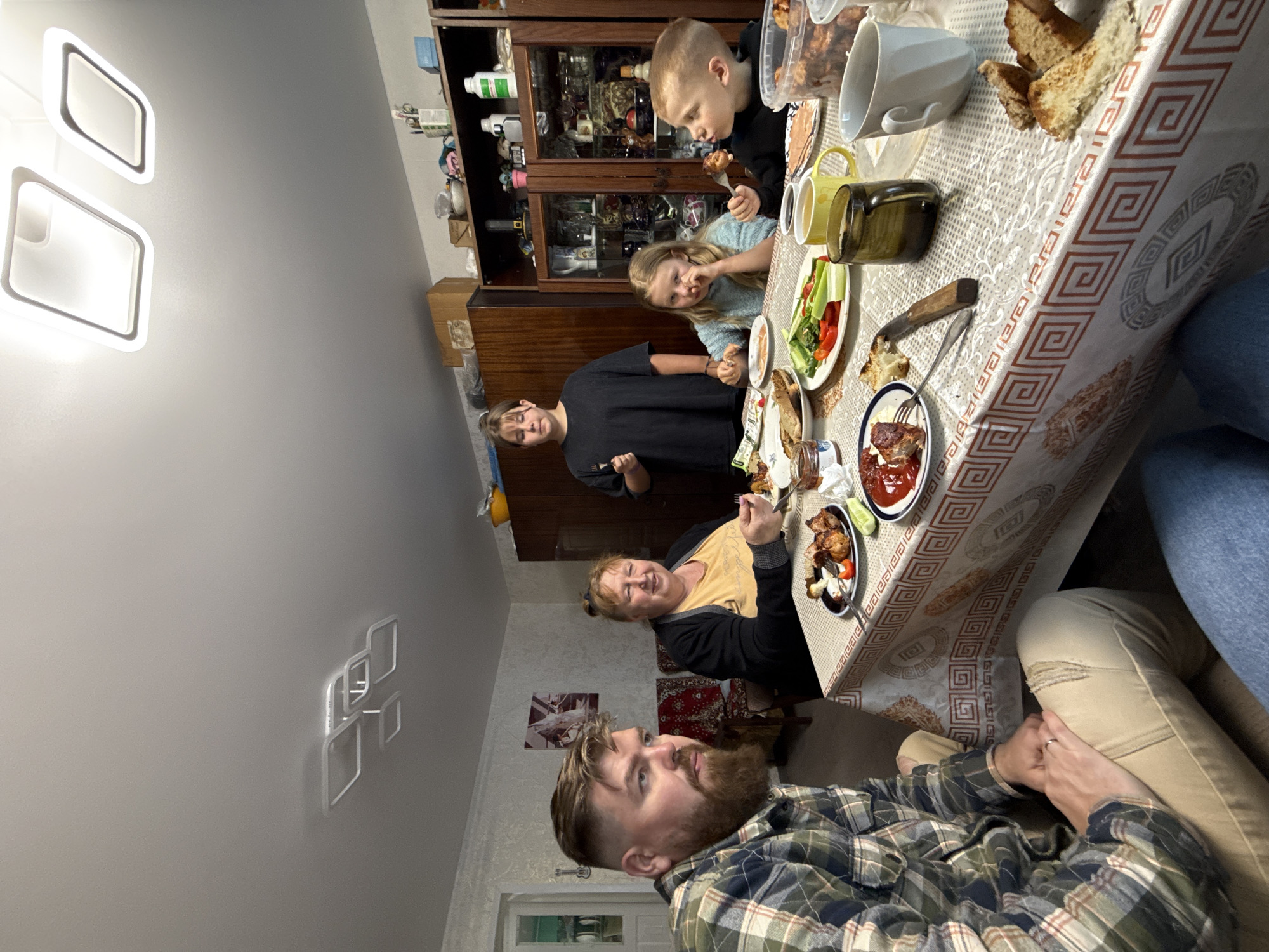 The image captures an indoor family gathering around a dining table in what appears to be a home in Бронница, Russia. The scene is brightly lit by modern, geometric ceiling lights and other artificial sources, suggesting it is evening or nighttime.
In the foreground, a man with a beard and a plaid shirt is seated, looking towards the right side of the frame. His hands are clasped, and part of his legs, wearing beige pants, are visible, along with another person's legs in blue jeans beside him.
Around a rectangular table covered with a patterned tablecloth featuring a Greek key-like design, several individuals are present. An older woman with short hair and a yellow top, overlaid with a black cardigan, smiles broadly as she eats, looking towards the bearded man. To her left, a younger woman or teenager in a black t-shirt stands behind the table. Further down the table, two children are seated: a boy in a black hoodie is eating with a fork, and a girl in a light blue top looks towards the foreground.
The table is laden with food, including plates of what looks like roasted meat (possibly shashlik), fresh vegetables (cucumbers, tomatoes, bell peppers), bread, and a dish of ketchup or red sauce. Various cups (white, yellow) and a dark green bottle or jar are also on the table.
In the background, a large wooden cabinet with glass doors displays various glassware and bottles, along with other household items on shelves. The overall atmosphere appears warm, familial, and relaxed, with people enjoying a meal together. There is no visible text.