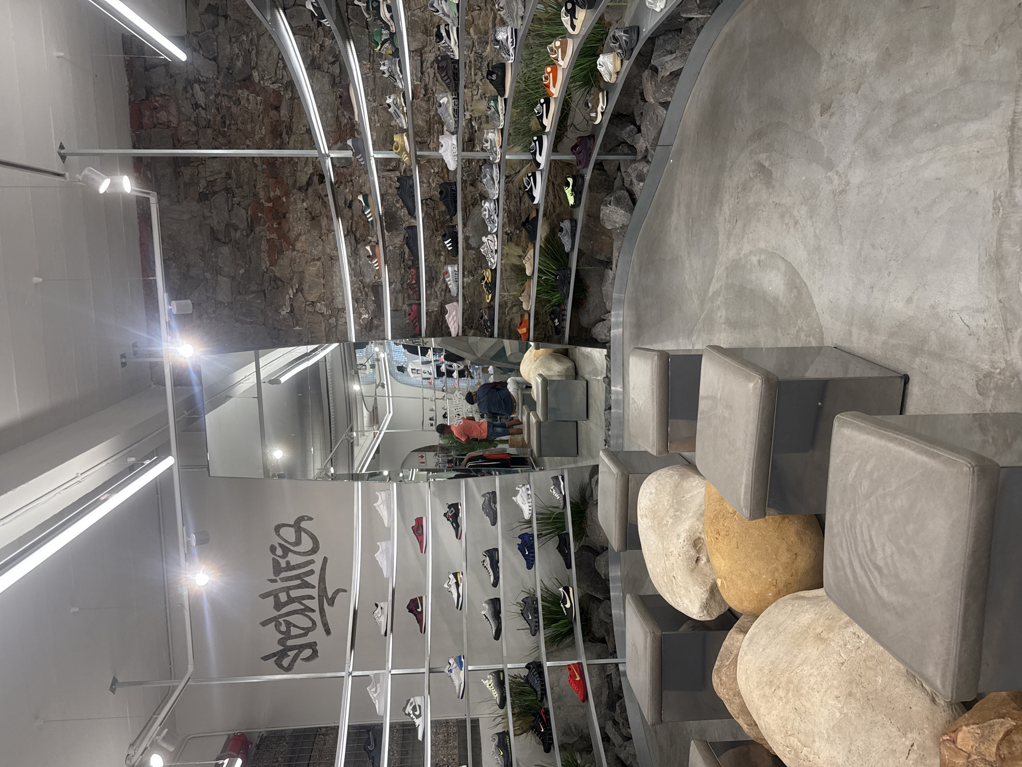The image displays the interior of a retail store, identified by the prominent "Shelflife" logo, located at 169 Loop Street, Cape Town, South Africa. The scene features a modern industrial design aesthetic with a polished light gray concrete floor. The right wall is composed of exposed rough-hewn stone and brickwork, while the left wall is painted a light gray, adorned with the dark gray "Shelflife" logo in a graffiti-style script.
Merchandise, primarily sneakers, is displayed on curved, multi-tiered metal shelves that line both walls, extending from near floor level upwards. Each shoe is individually presented, with the lower sections of the display incorporating dark, irregular rocks and scattered artificial green plants. The store is brightly illuminated by fluorescent tube lighting and spotlights mounted on the ceiling and along structural poles.
In the foreground and mid-ground, multiple square stools with light gray padded tops and reflective silver bases are arranged on the floor. These are interspersed with several large, natural boulders in off-white and earthy brown hues.
A large, tall mirror on the left side of the background reflects an additional section of the store. Within this reflection, two adult males are visible. One man, dressed in a red short-sleeved shirt and blue shorts, stands near a clothing rack. The second man, wearing a dark blue top and light trousers, is seated on a light-colored stool, his back mostly to the viewer. No direct interaction is depicted between these individuals or with the immediate foreground.