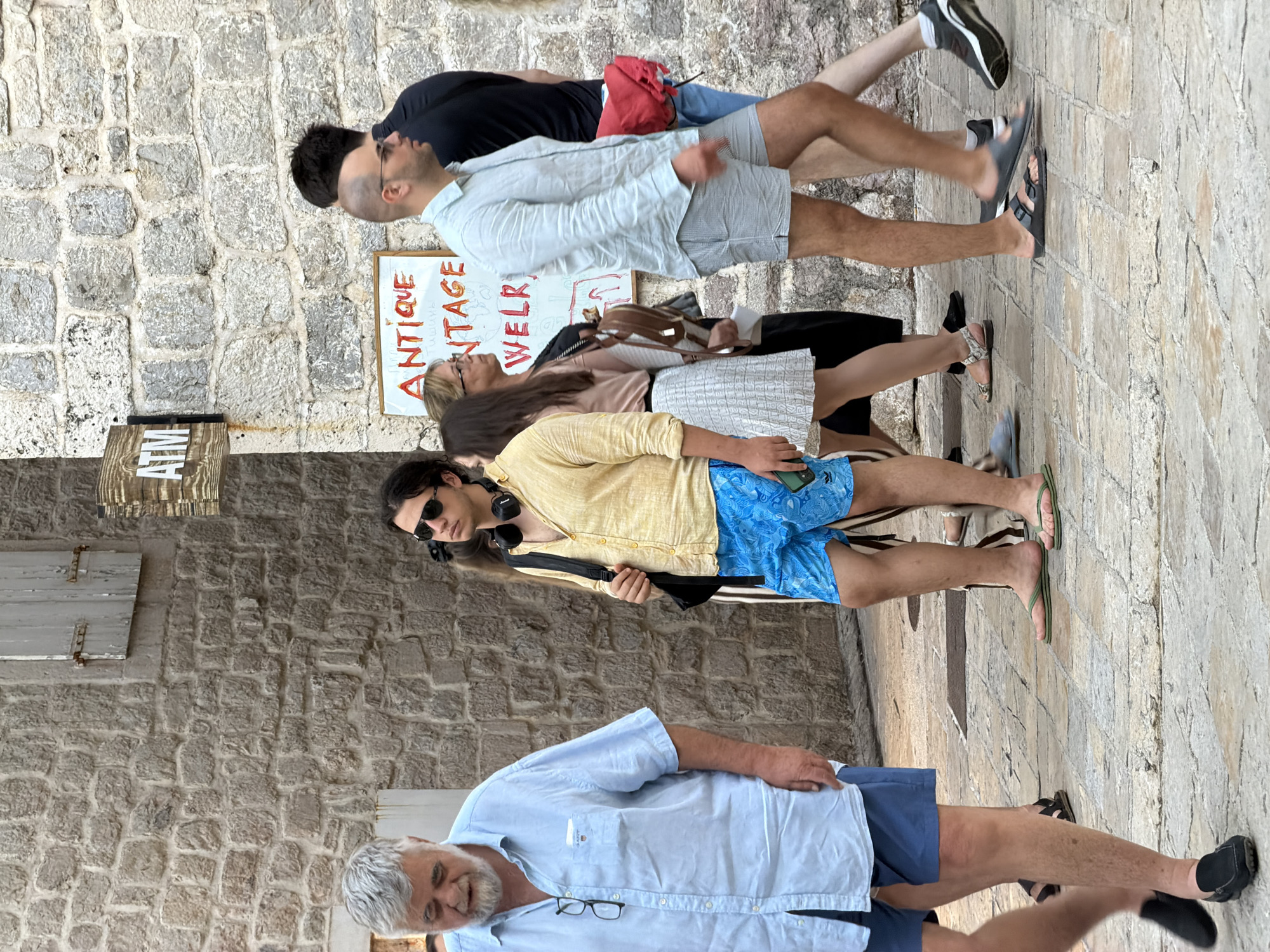 A group of people walk along a stone-paved street in the historic Old Town of Budva, Montenegro. The buildings are constructed of rough-hewn stone blocks, with some featuring wooden shutters. In the background, an ATM sign is visible on a stone wall. Several individuals are walking in various directions, some appearing to be tourists. One man in the foreground wears a light blue short-sleeved button-down shirt and blue shorts. Another man, slightly behind and to his right, is dressed in a yellow short-sleeved shirt and bright blue patterned shorts, with headphones around his neck. Further to the right, a man in a light blue shirt and grey shorts walks away from the camera. A woman with blonde hair wearing glasses and a light-colored skirt is also visible. The pavement is made of evenly-cut stone tiles.