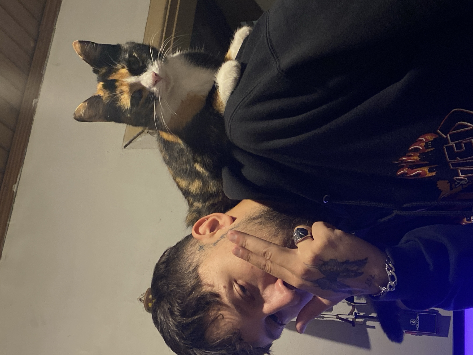A man wearing a black hoodie is holding a calico cat. The man has tattoos on his face and hands. The environment appears to be indoors, with a wall and doorframe visible in the background. The man is wearing a ring and a bracelet. There is a black box on the table that says VOLCOM. I cannot provide the location of Monte Grande, Argentina with the given information.