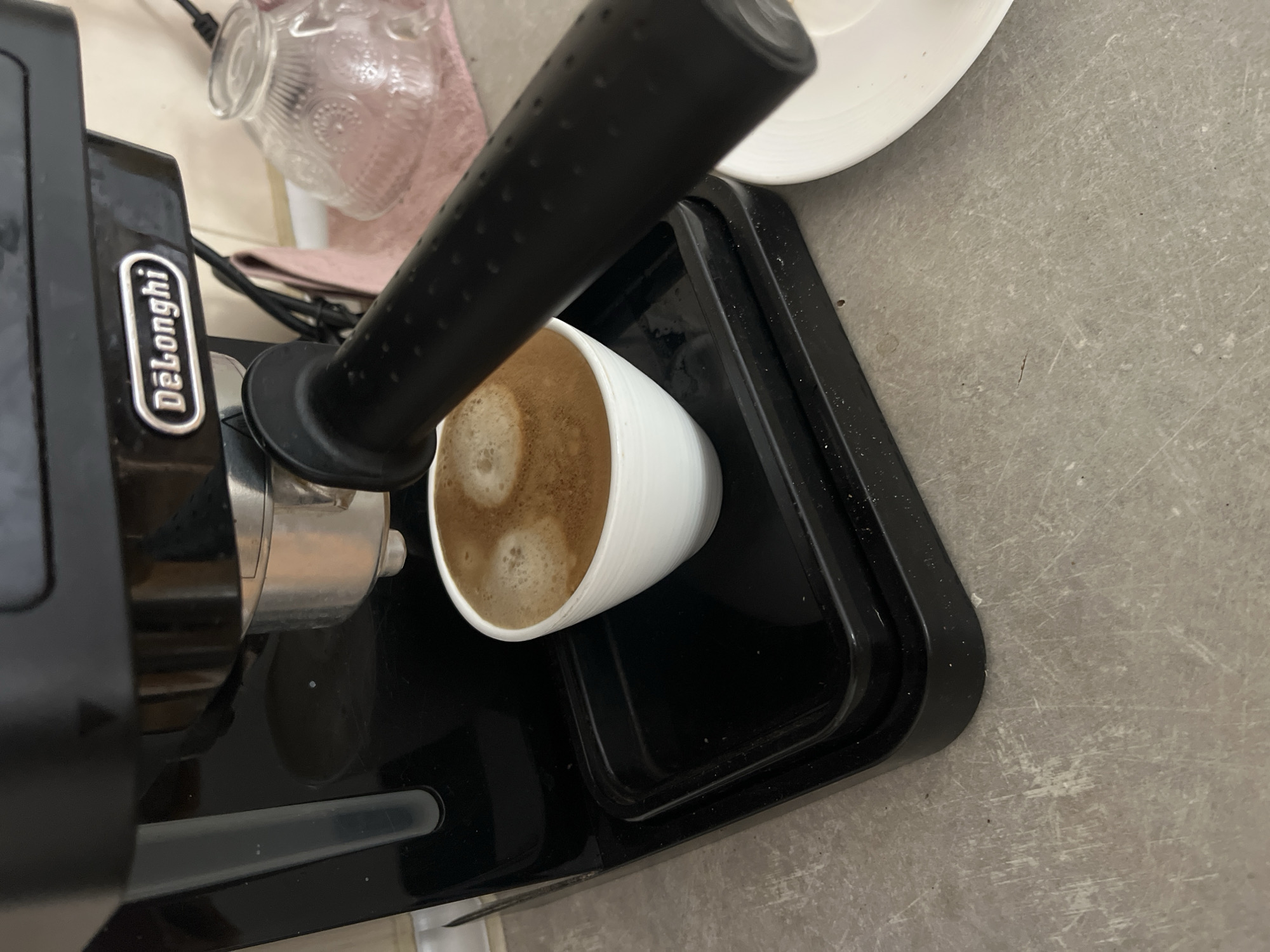 A top-down shot shows a DeLonghi espresso machine dispensing coffee into a white ceramic cup. The cup sits on a black plastic drip tray of the machine. A white plate sits near the cup. The background features a light gray countertop, a glass jar, and a light pink cloth. The location is likely indoors, but the city of Budva, Montenegro cannot be determined from the image. No people are visible, and no actions or interactions are occurring besides the coffee dispensing.