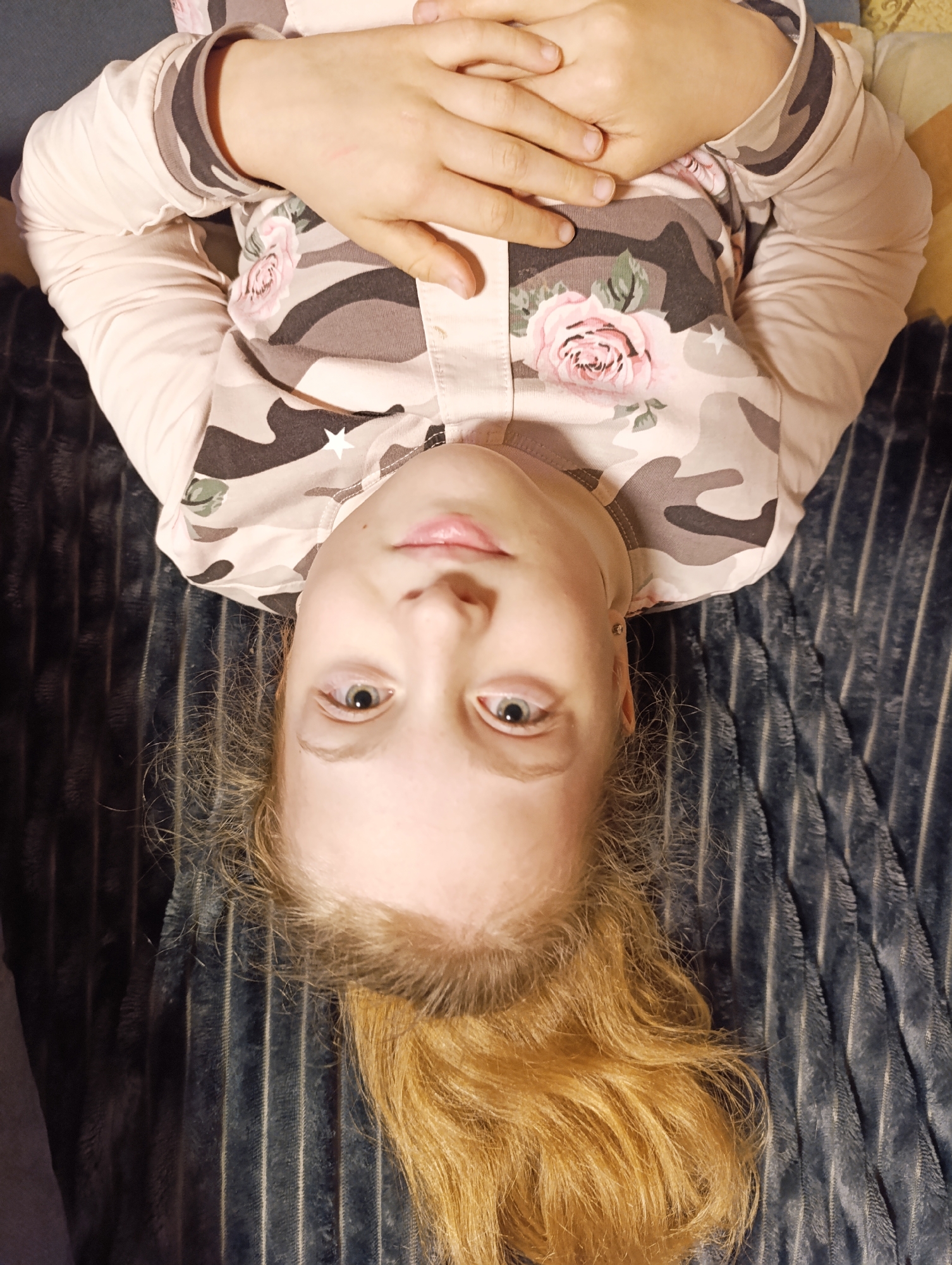 A young person with blonde hair, tied back in a low ponytail, is lying on a blue blanket. The person is wearing a long-sleeved shirt with a camouflage pattern featuring pink roses and white stars. Their hands are folded across their chest. The location is Unknown, Unknown.